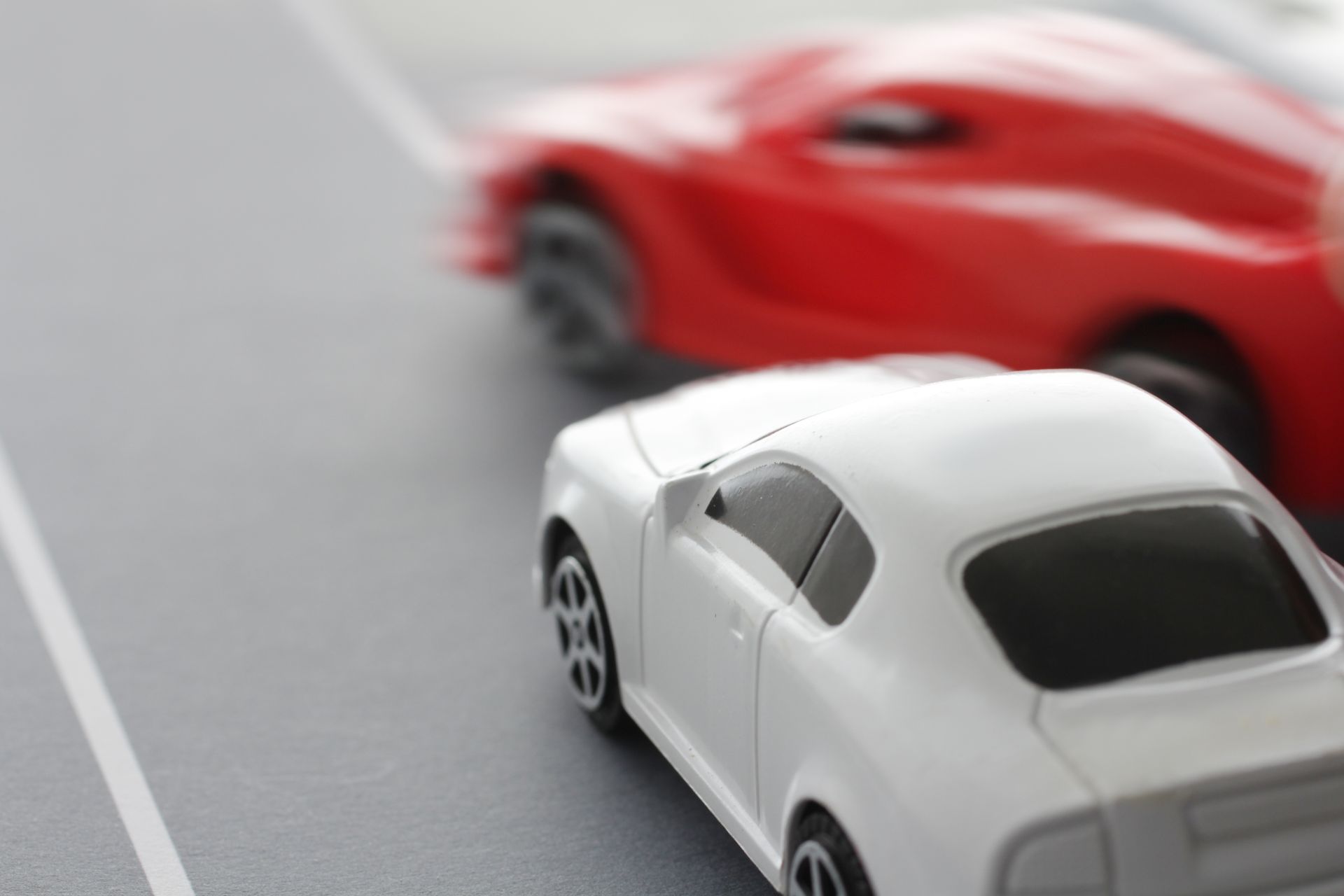White toy car in the foreground with a blurred red car passing on a road