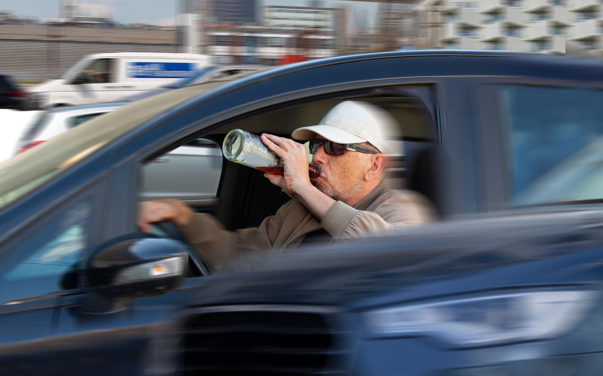 Driver in white cap drinking from a bottle while driving a blue car on a city street.