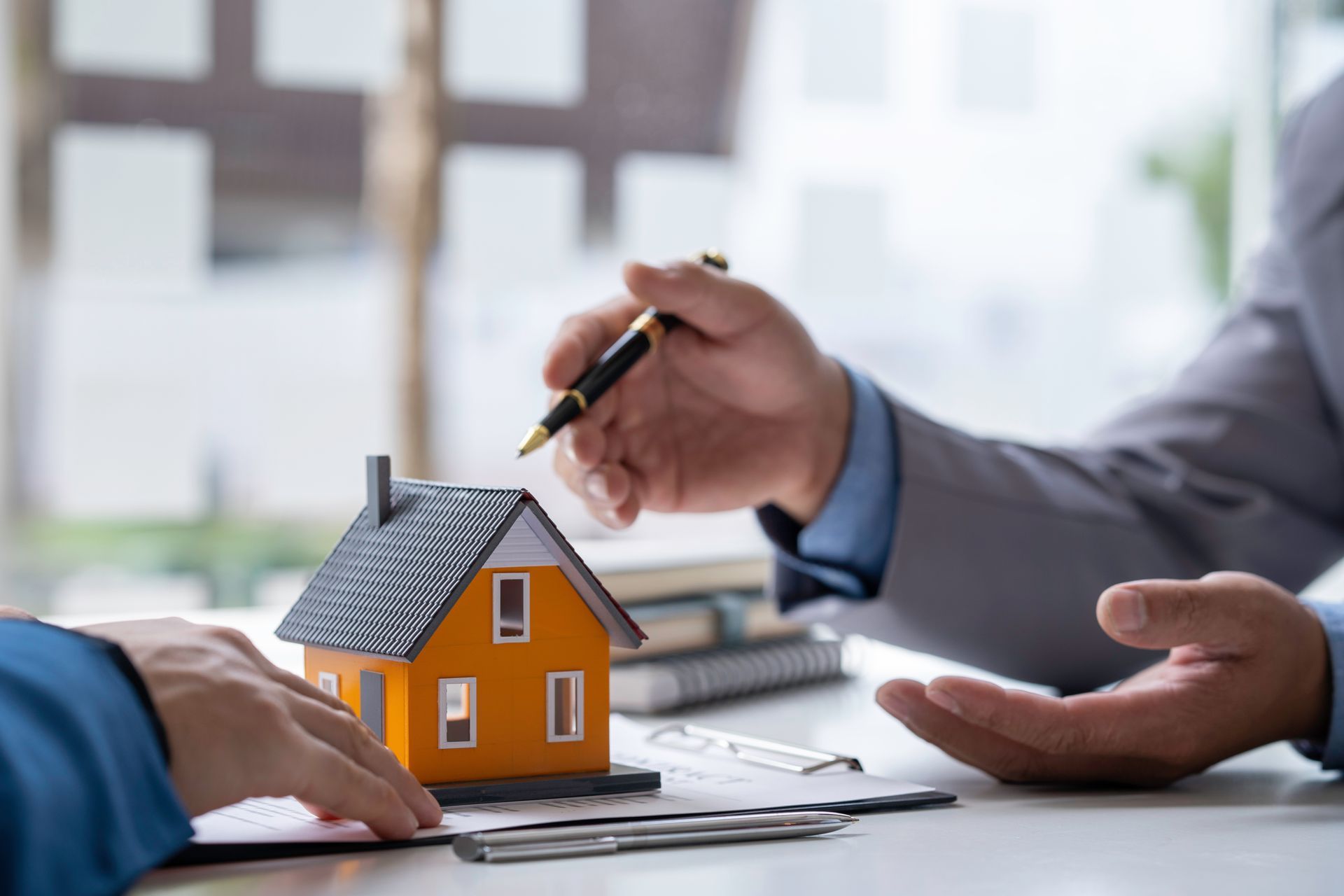 Hands discussing a small house model on a desk with a pen, suggesting a real estate meeting