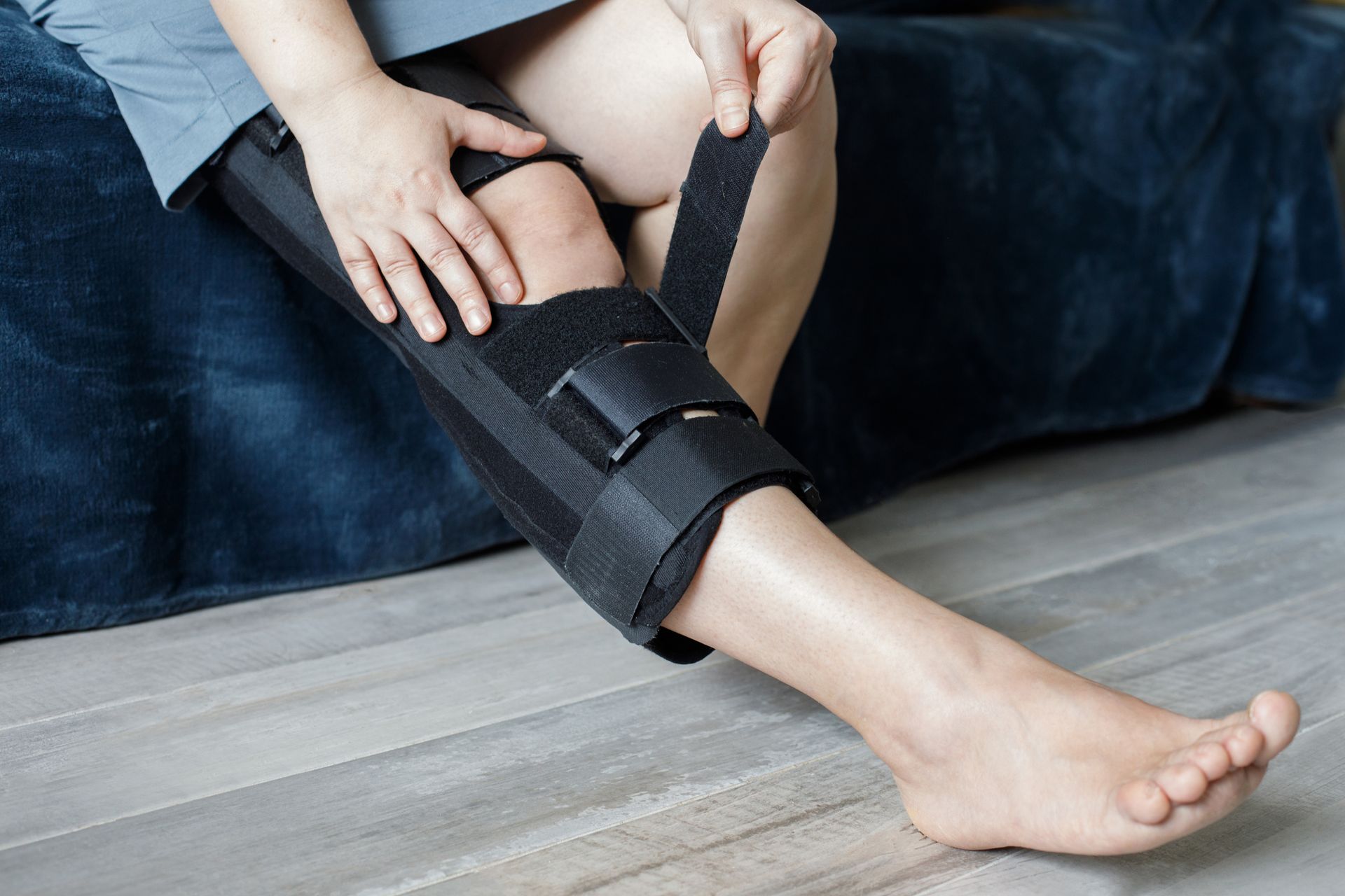 Person fastening a black leg brace on a lower leg while sitting on the floor.