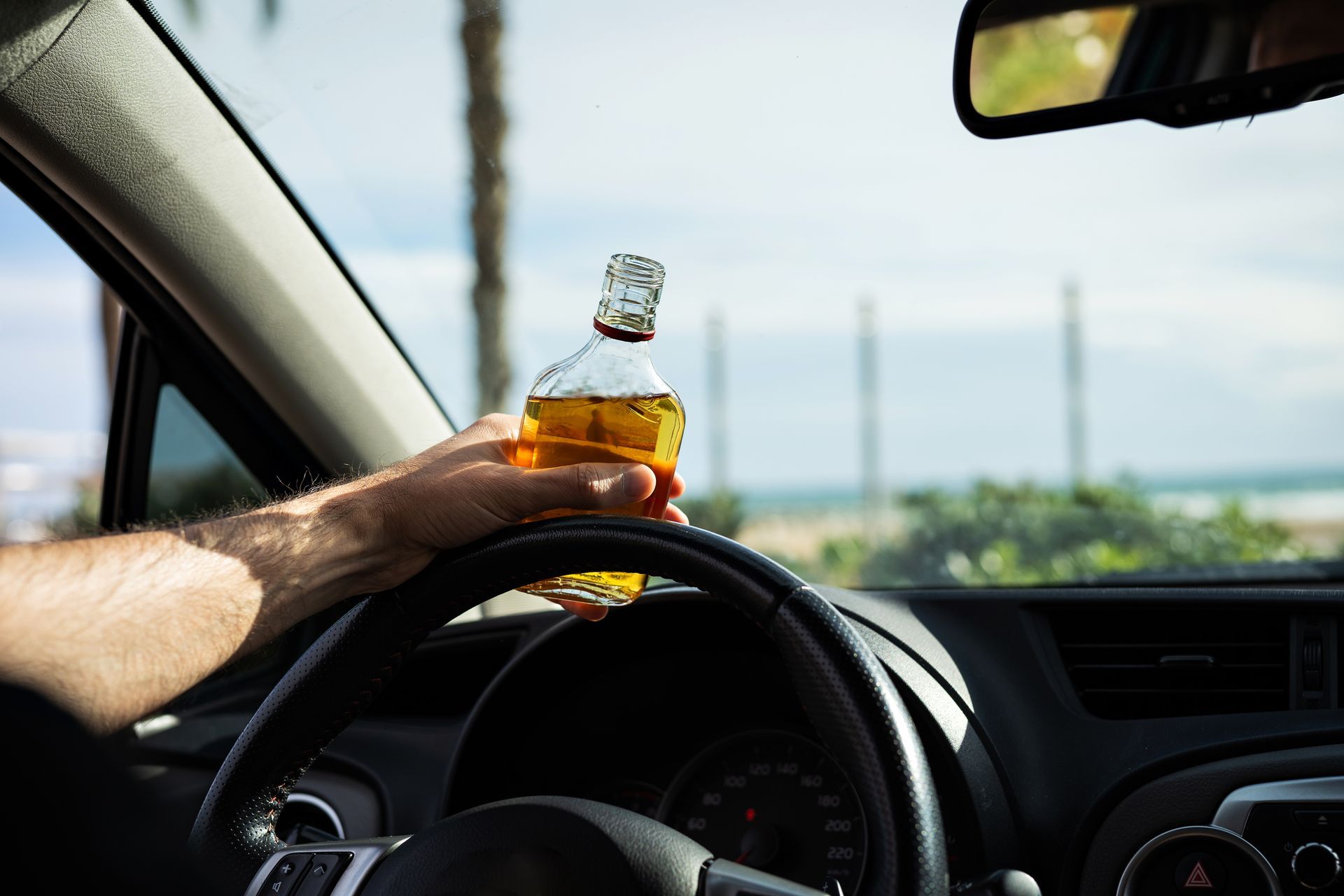 Hand holding a whiskey bottle over a steering wheel inside a car
