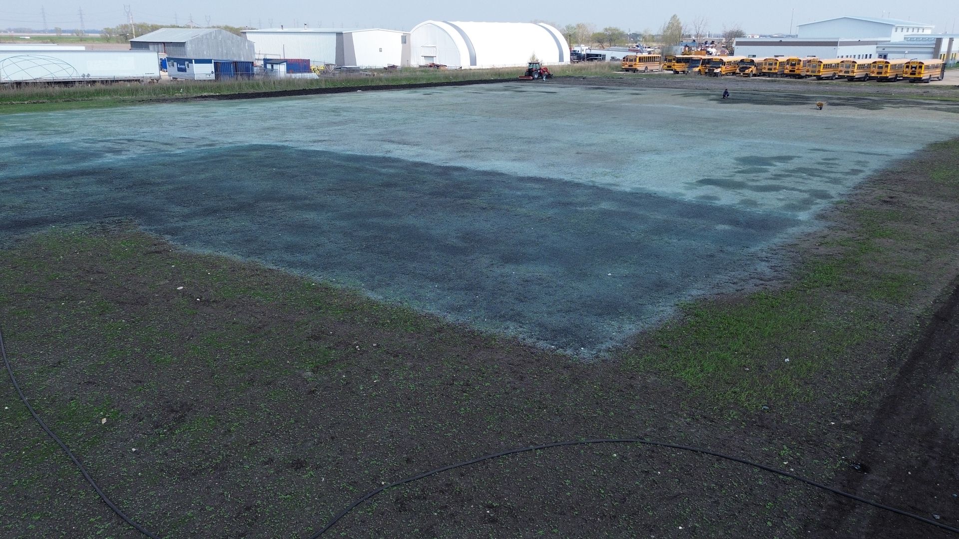 Grassy field with patches of dark material, with industrial buildings in the background on a sunny day.