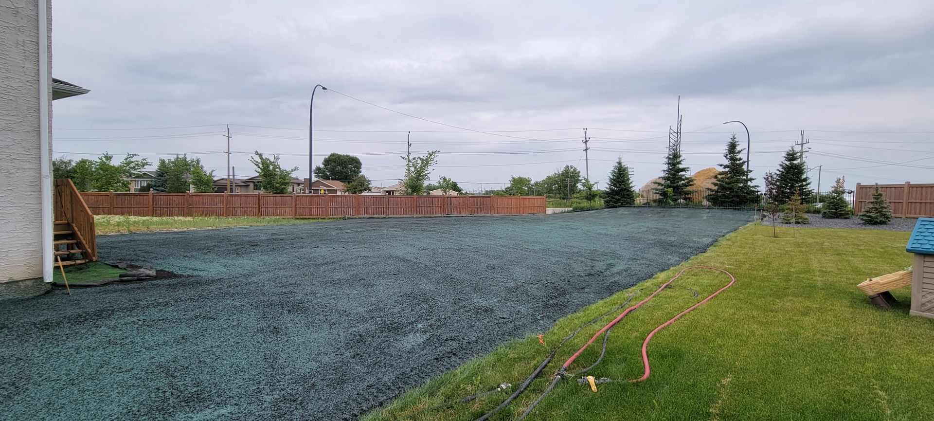 Backyard with a dark green ground cover, green grass, a wooden fence, and a cloudy sky.