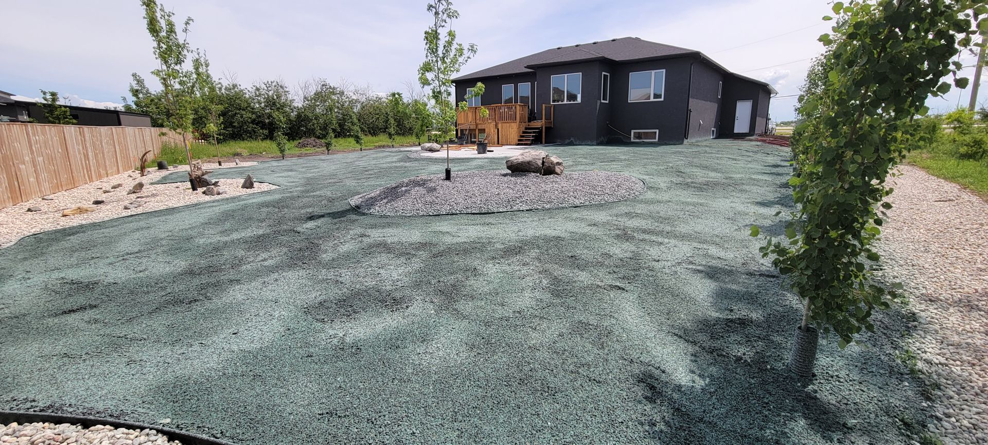 A house with dark siding and a wooden deck, surrounded by gravel and artificial turf.