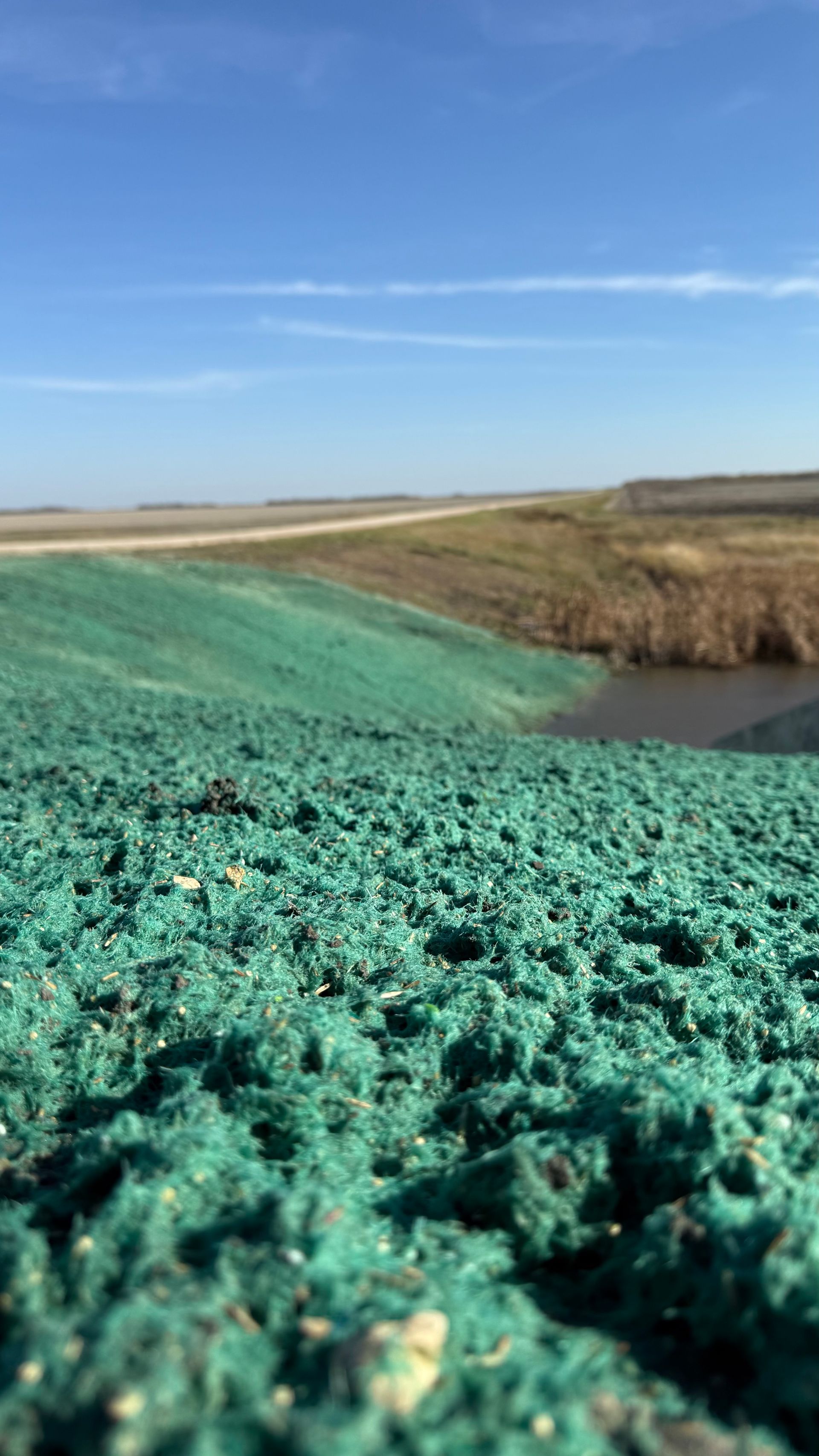 Green material covers a slope; blue sky in background, a road in the distance.