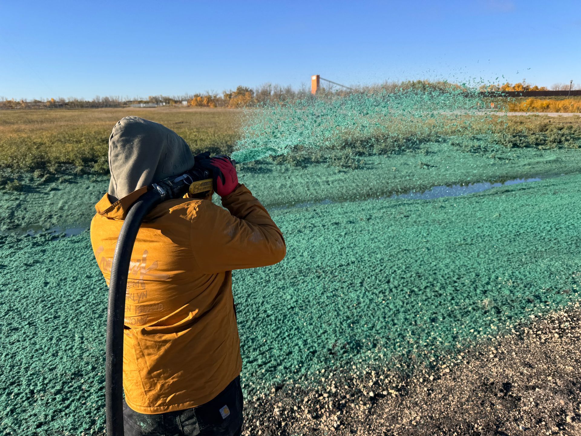 Person spraying green liquid on field, wearing protective gear, blue sky, and farmland in background.
