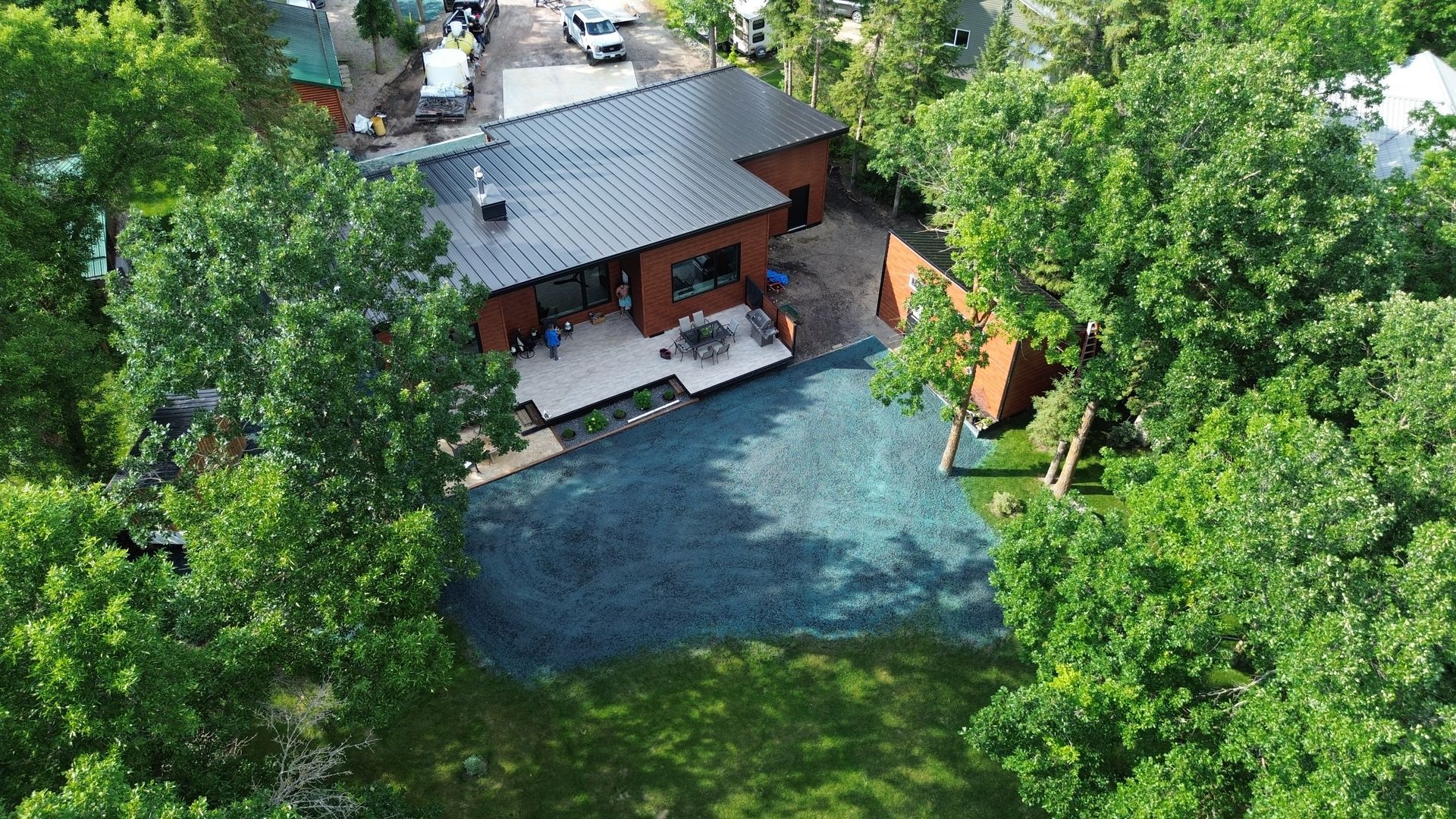 Aerial view of a modern brown house with a black roof and deck, surrounded by green trees and grass.