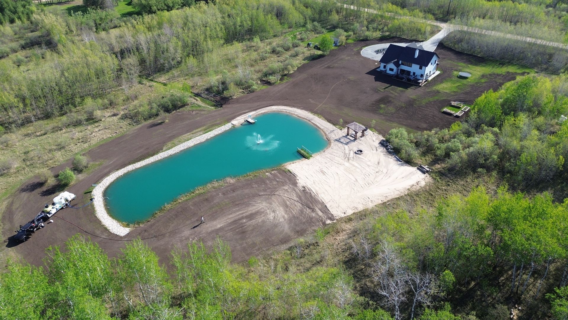 Aerial view of a teal-colored pond with a fountain, a house, and surrounding greenery.