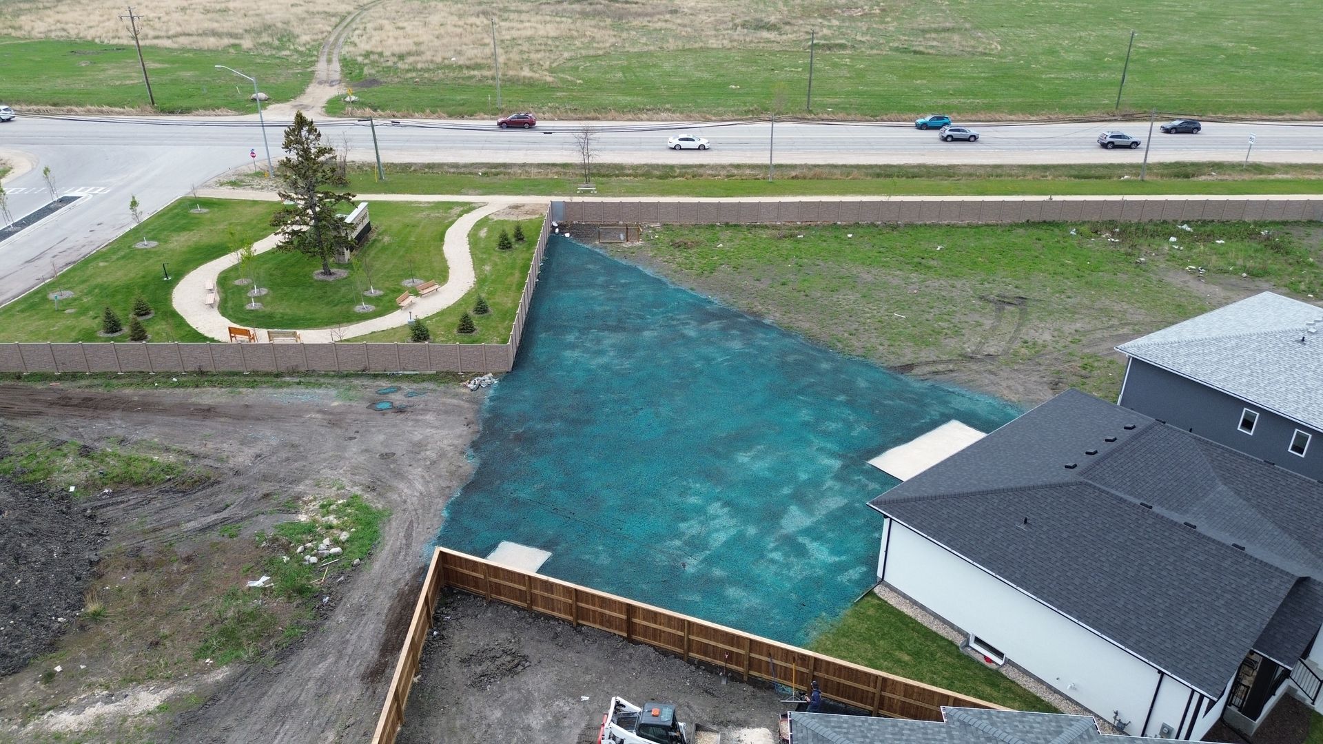 Aerial view of new construction: houses, freshly seeded lawn with green netting, road with cars, and a small park area.