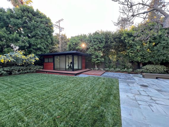Backyard garden with lawn, stone patio, and a small red shed surrounded by trees and hedges