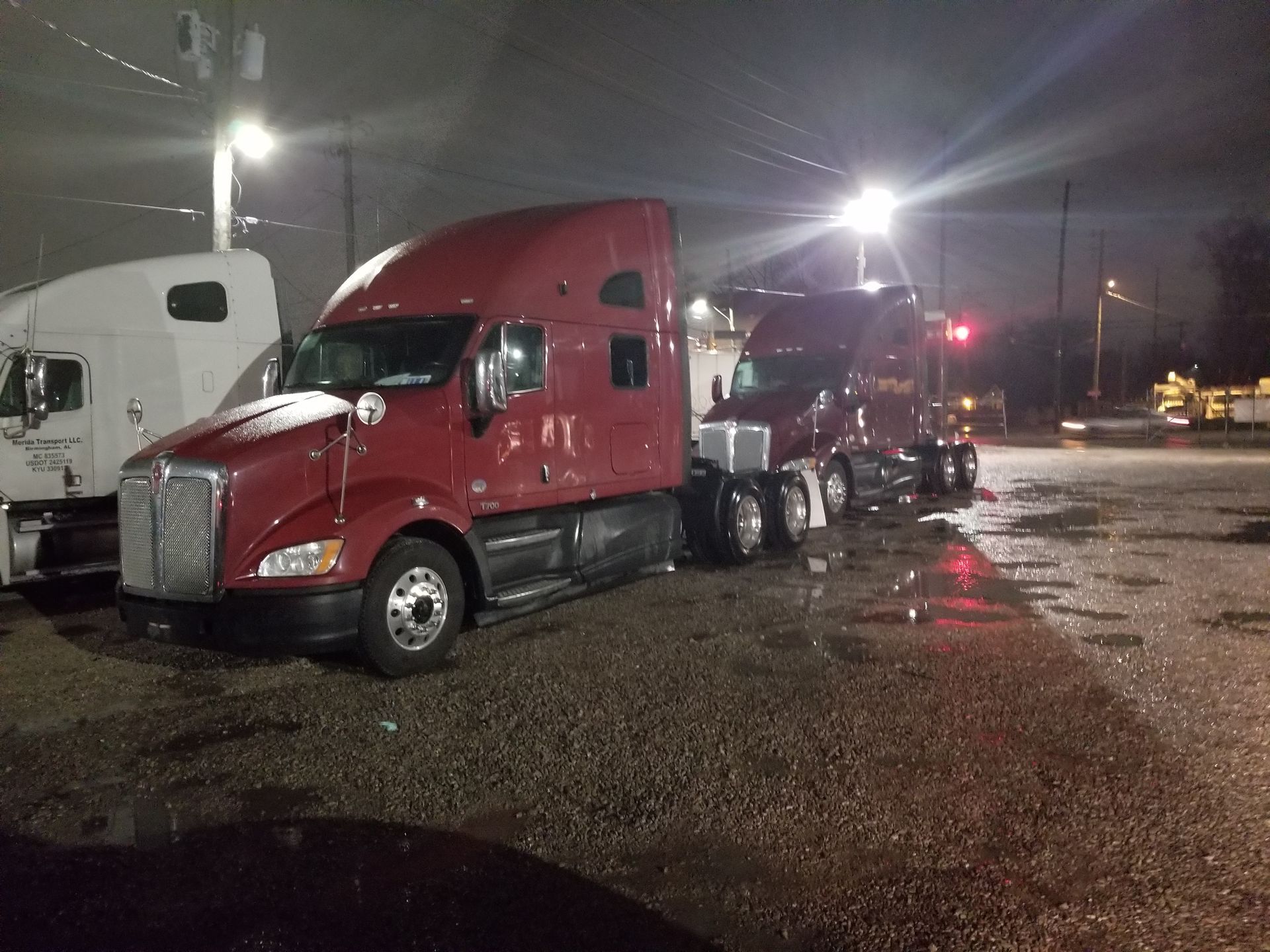 A row of semi trucks are parked in a parking lot at night.