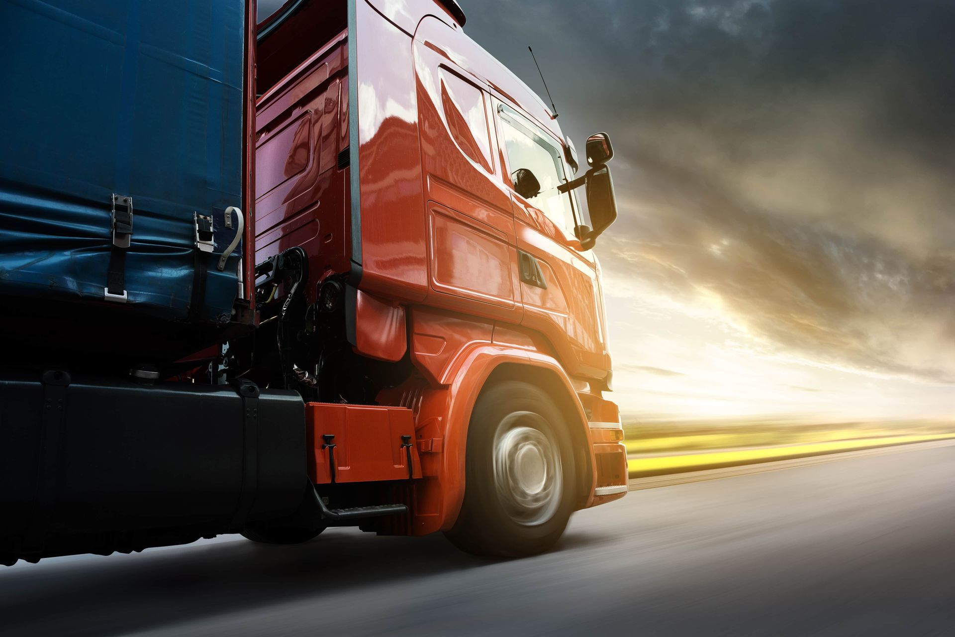 A red semi truck is driving down a highway at sunset.