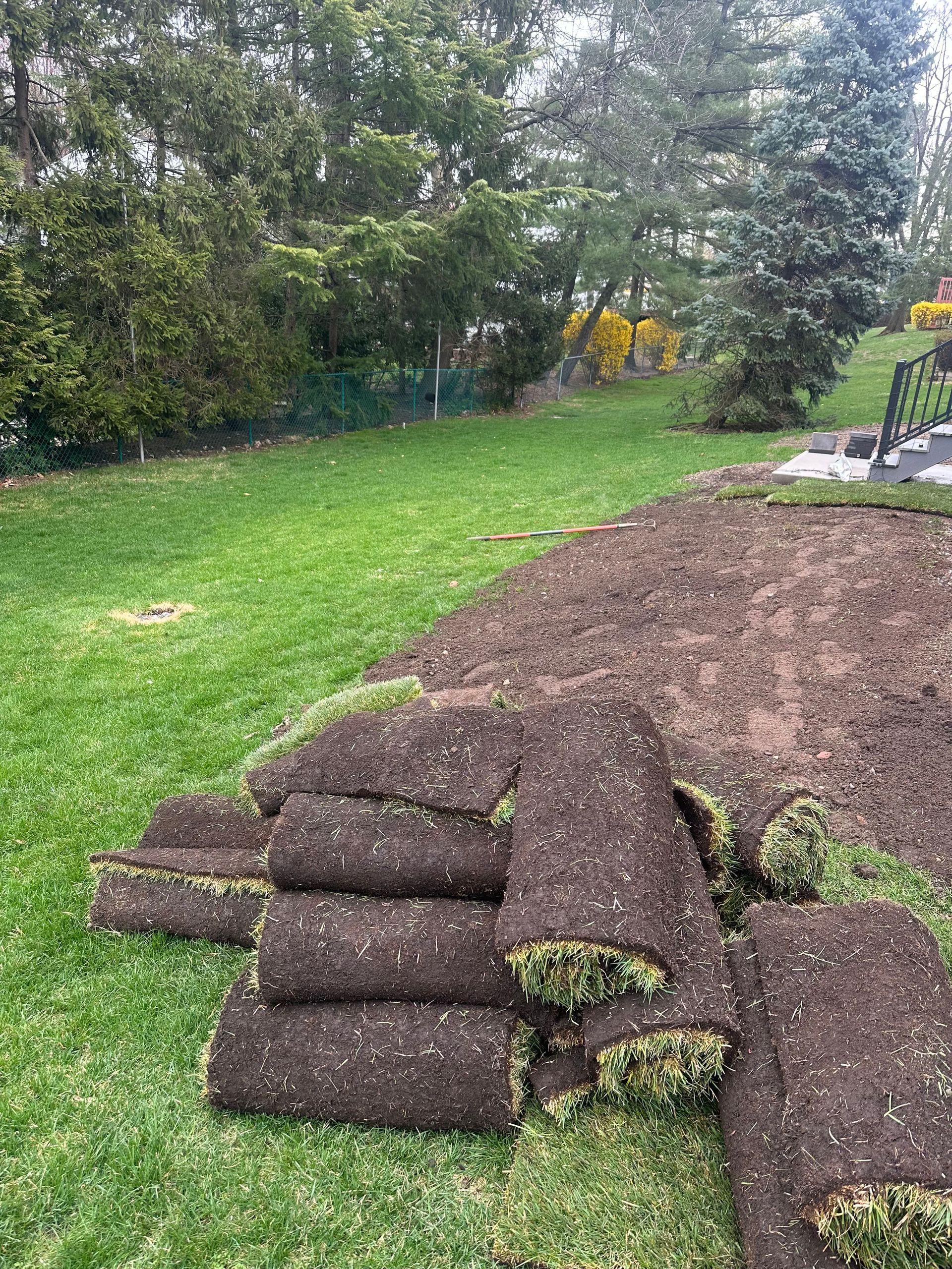 Stacked rolls of brown landscaping fabric beside a tilled garden bed in a grassy backyard.
