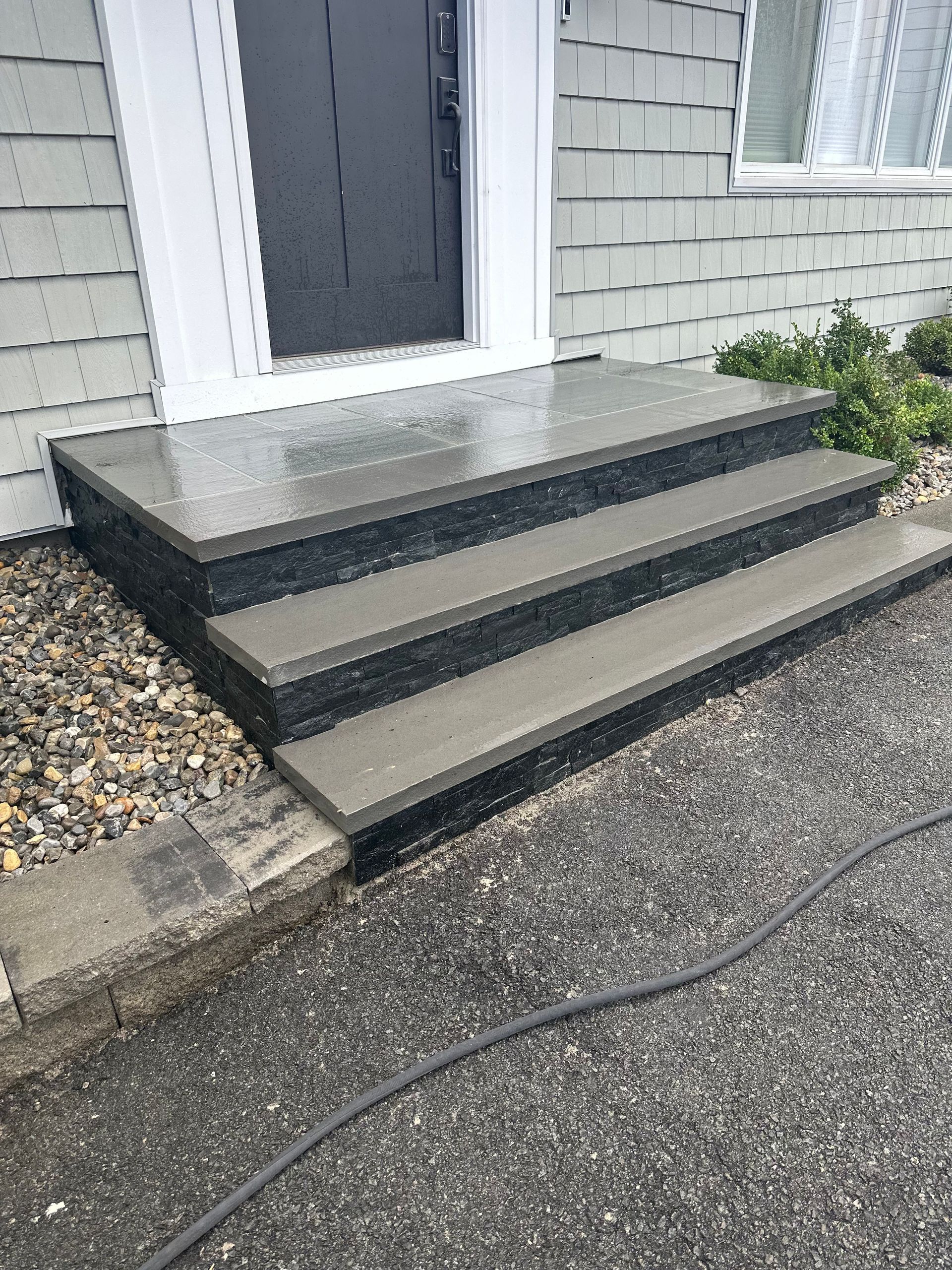 Front steps with three dark concrete risers leading to a house entry door and gravel walkway