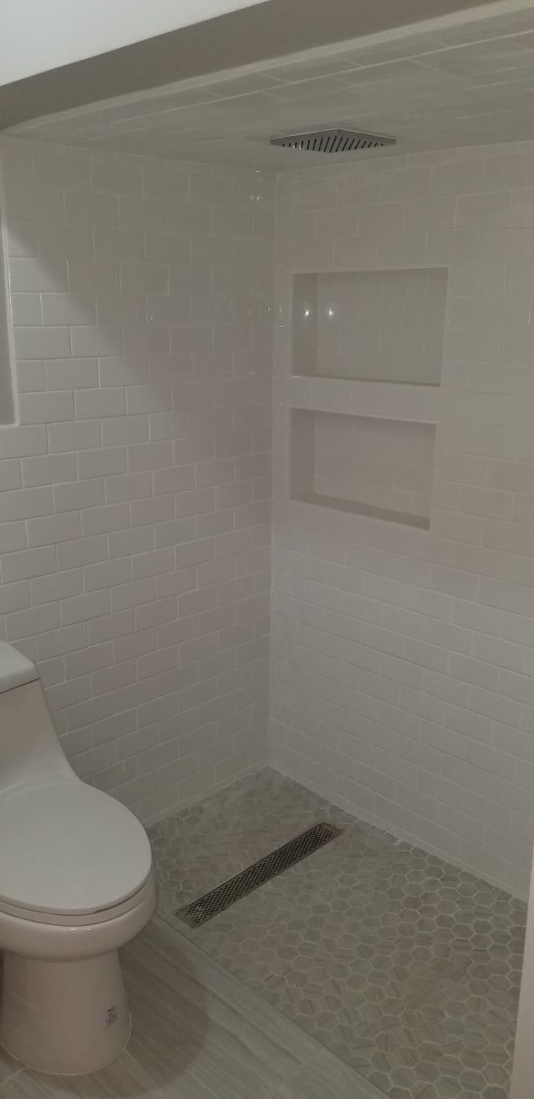 White tiled bathroom with toilet, shower, wall shelves, and floor drain