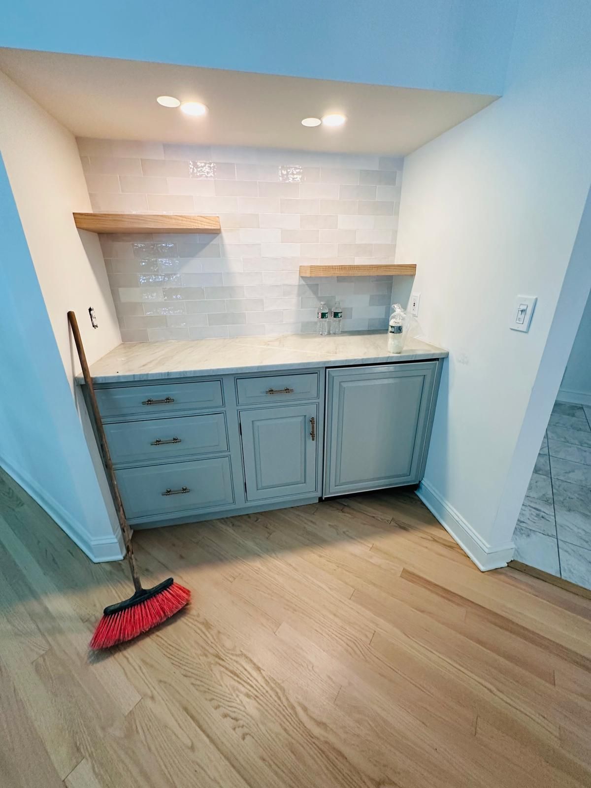 Small kitchen alcove with blue cabinets, white tile backsplash, wood floor, and broom on the floor