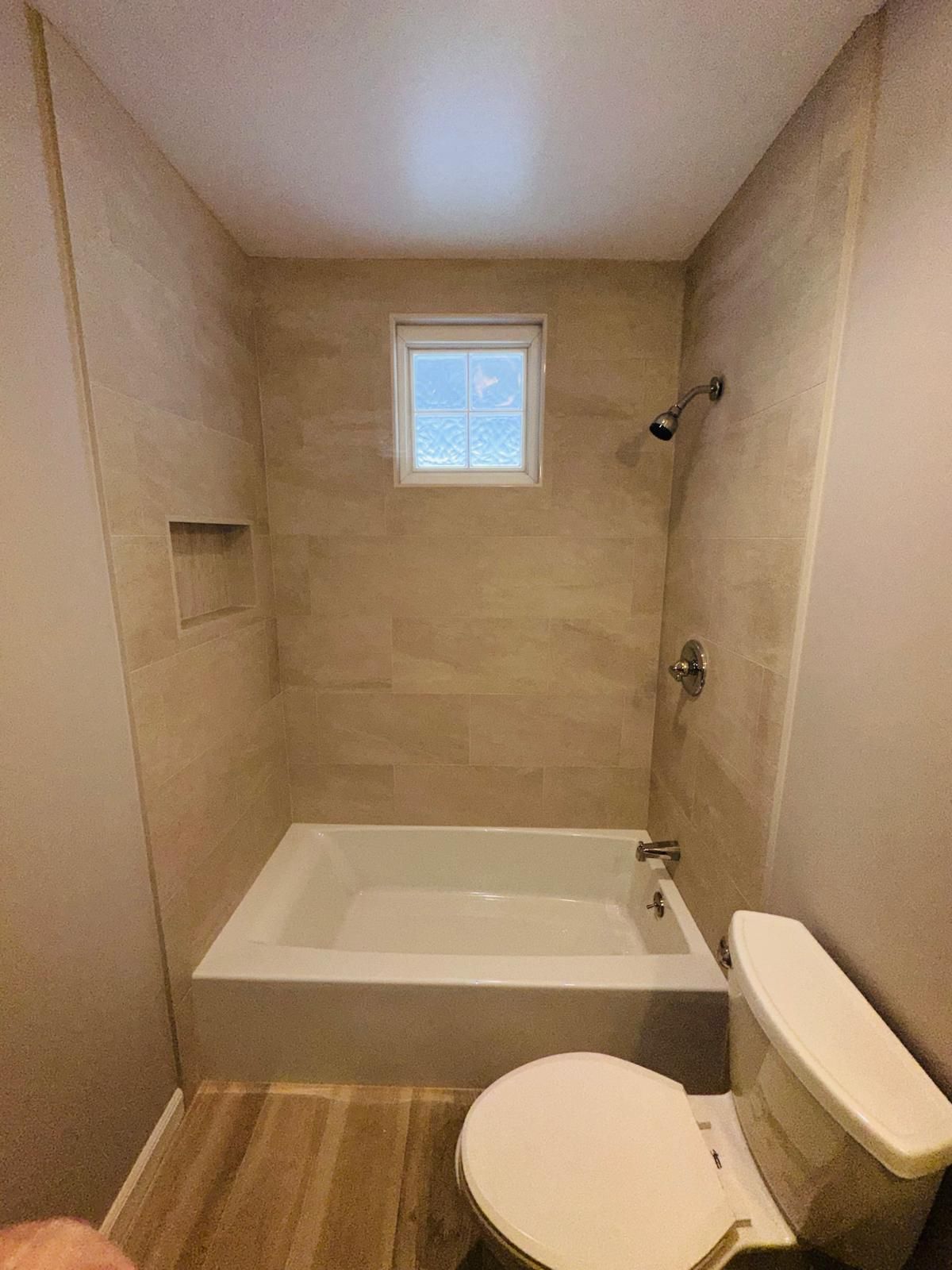 Small beige bathroom with a bathtub, toilet, showerhead, and a small window.