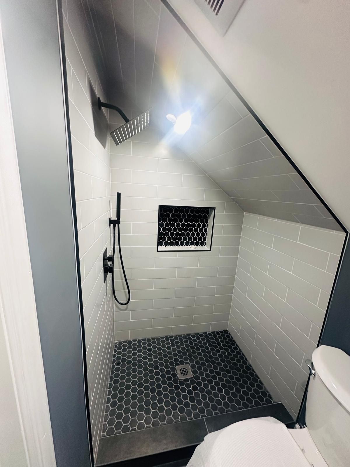 Compact tiled shower stall with handheld shower, small window, and sloped ceiling.