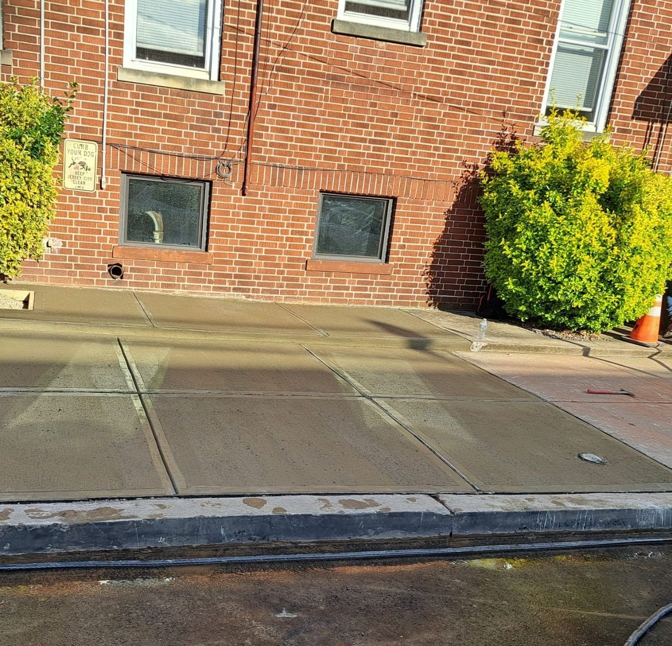 Brick building beside sidewalk and curb, with small basement windows and a green shrub on the right.