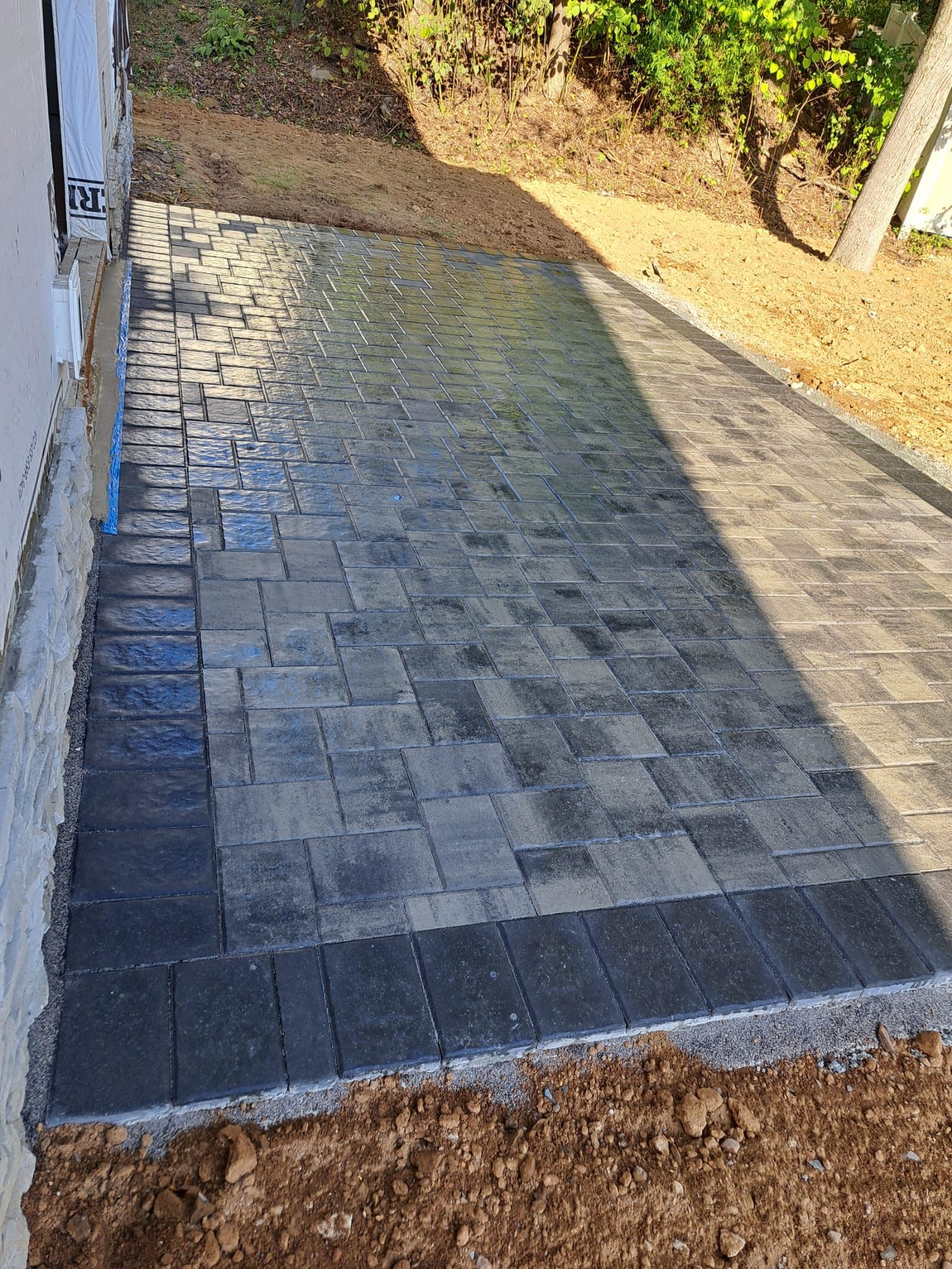 New gray paver patio beside a house, with soil in the foreground and a sloped yard beyond