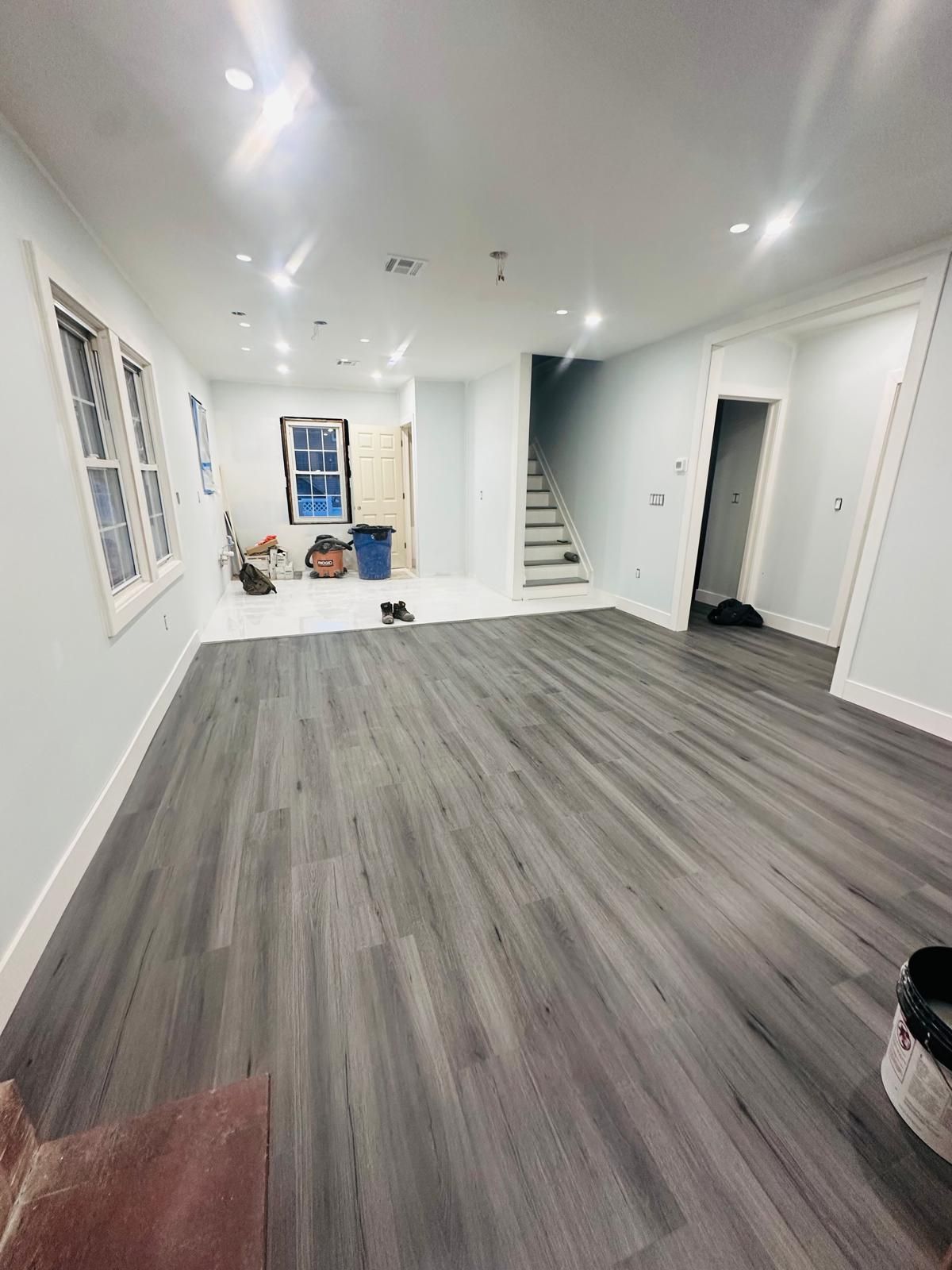 Empty bright living room with gray wood floors, white walls, and a staircase at the back.