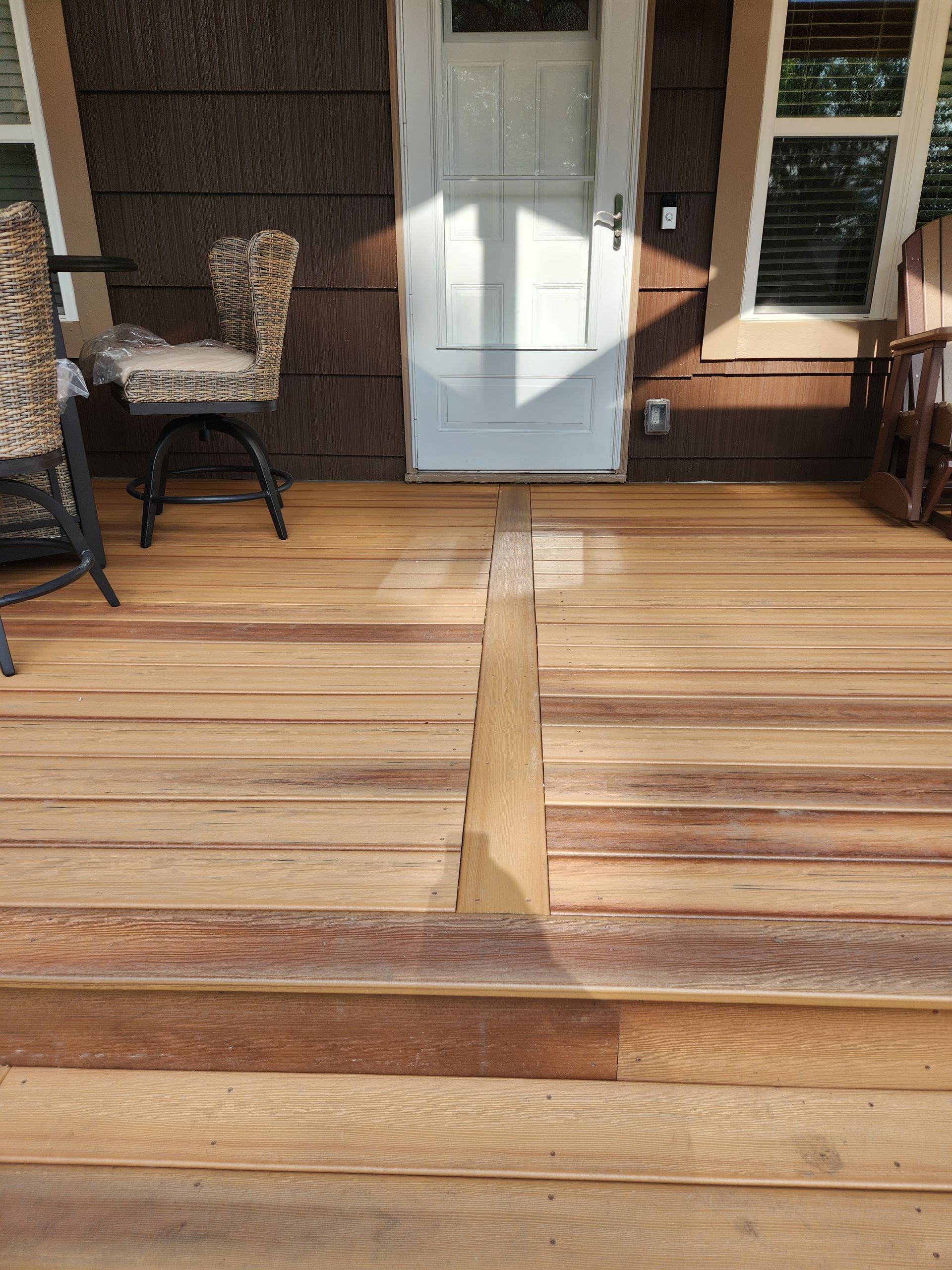 Wooden porch deck with a white door, wicker chair, and a sunlit board repair in the center.