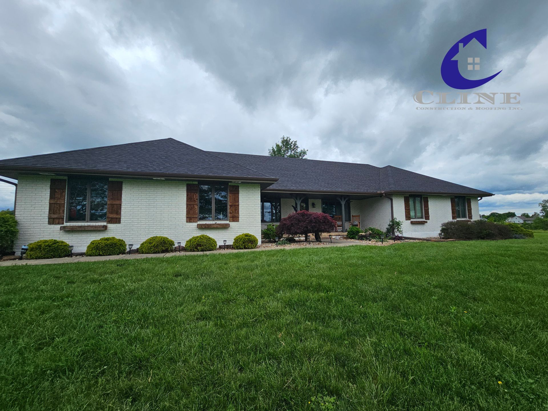 Single-story suburban house with dark roof, white brick exterior, and a green lawn under cloudy skies