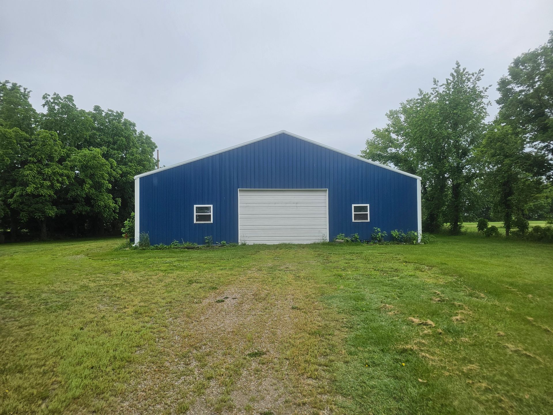 Blue metal barn with white garage door in a grassy field, framed by trees under an overcast sky