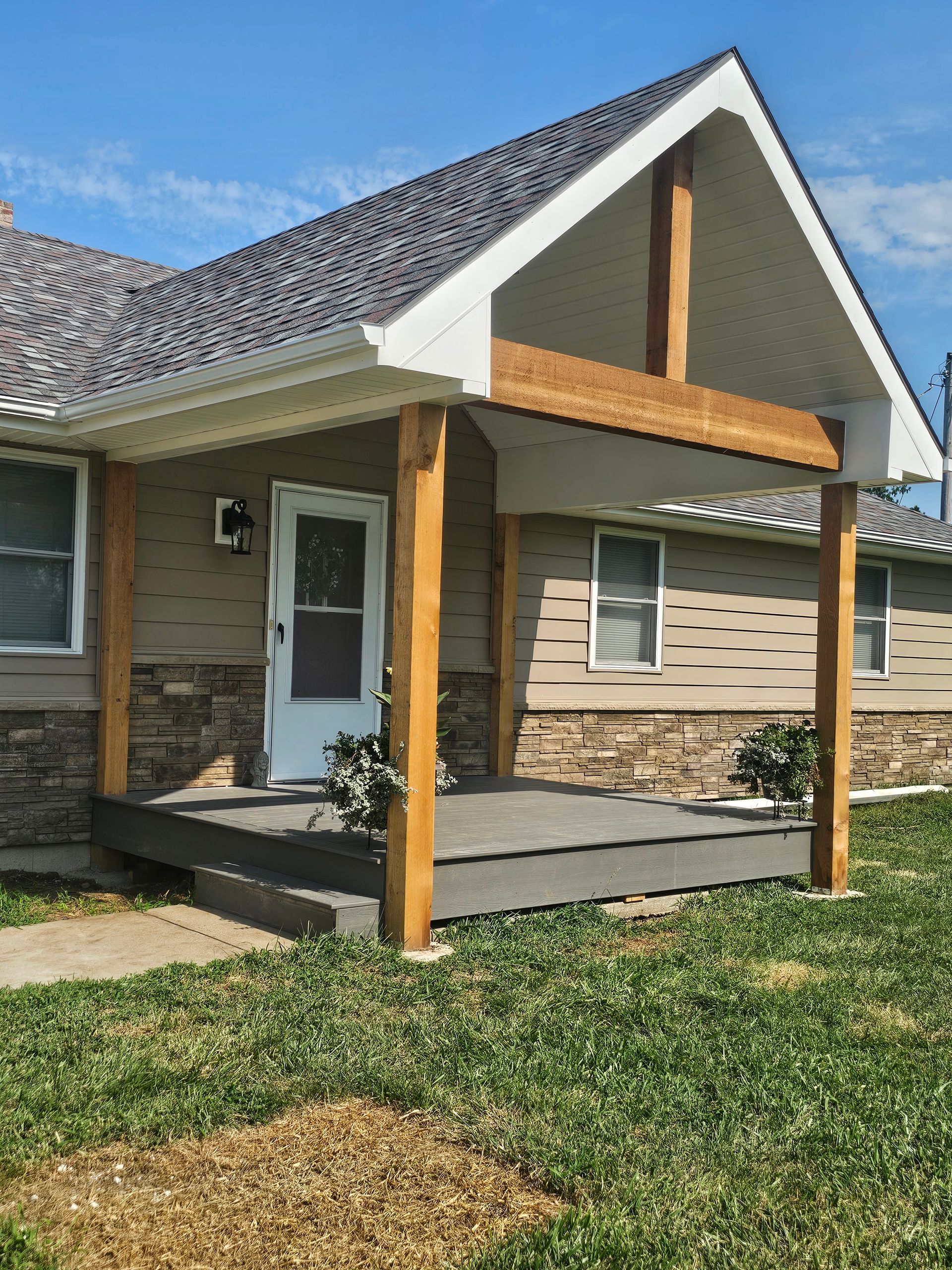 Front porch with wooden posts, stone facade, and white door on a suburban house under a clear blue sky