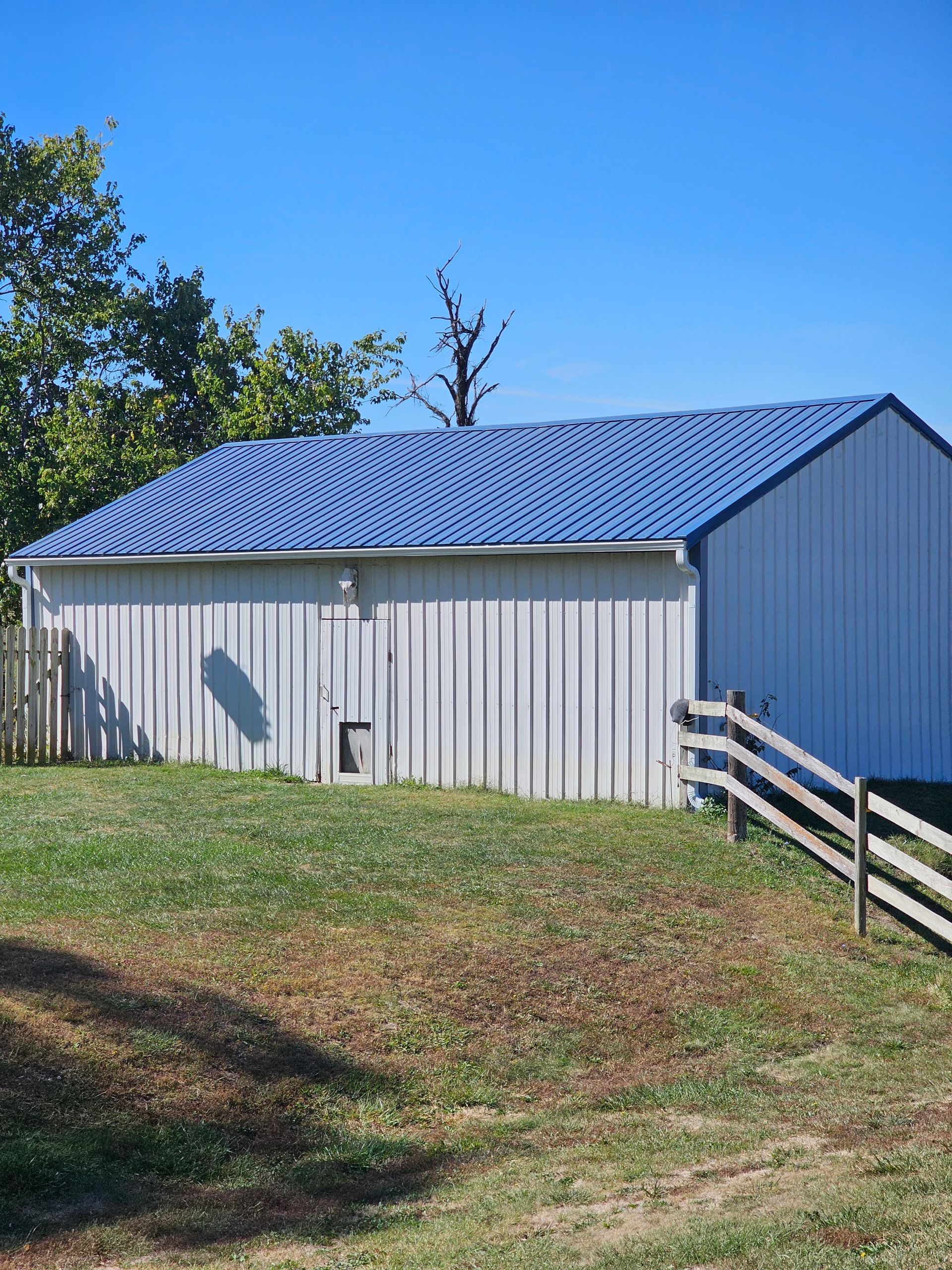 Small white shed with a blue metal roof beside a wooden fence on a sunny grassy hillside