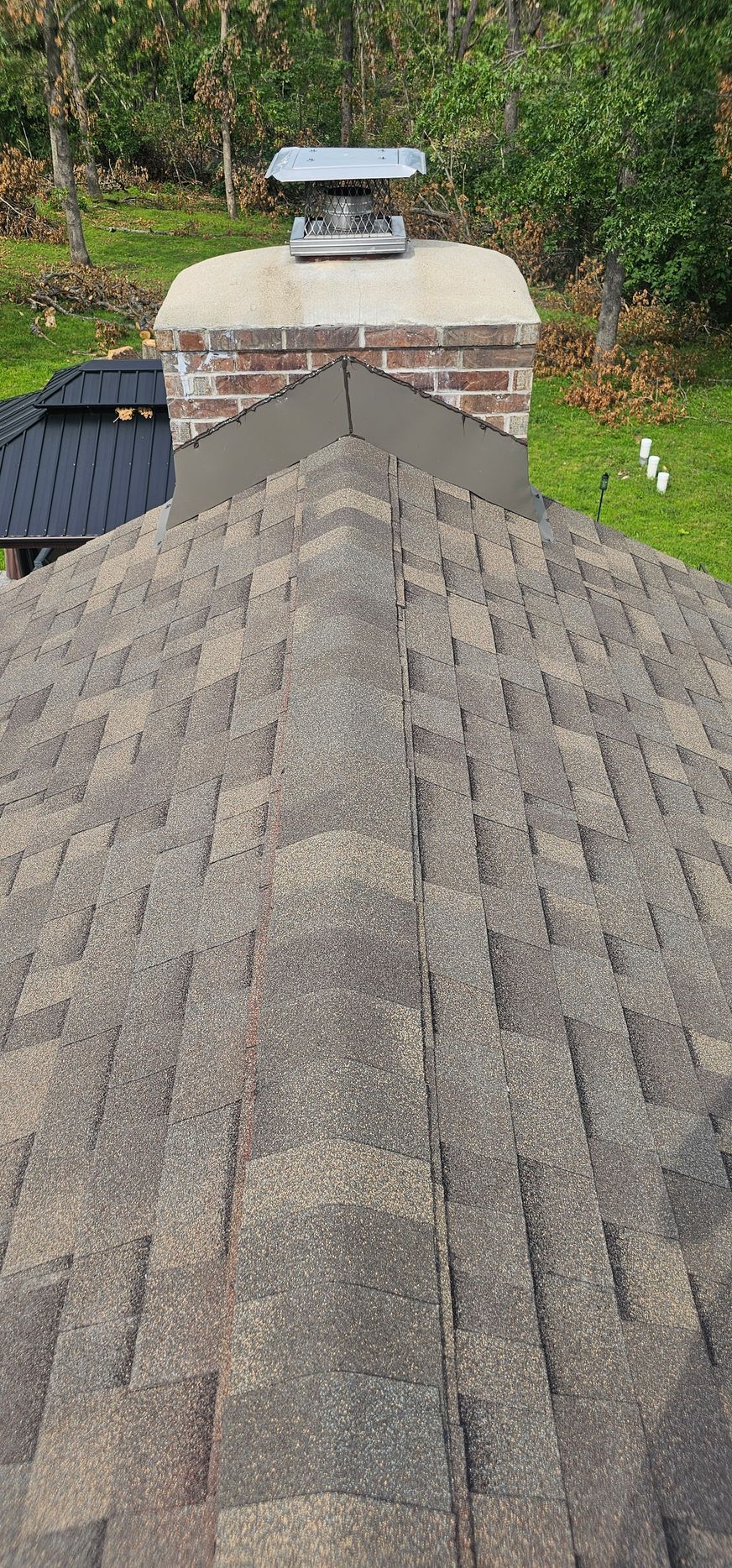 Shingled roof leading to a brick chimney with cap, backed by trees and greenery
