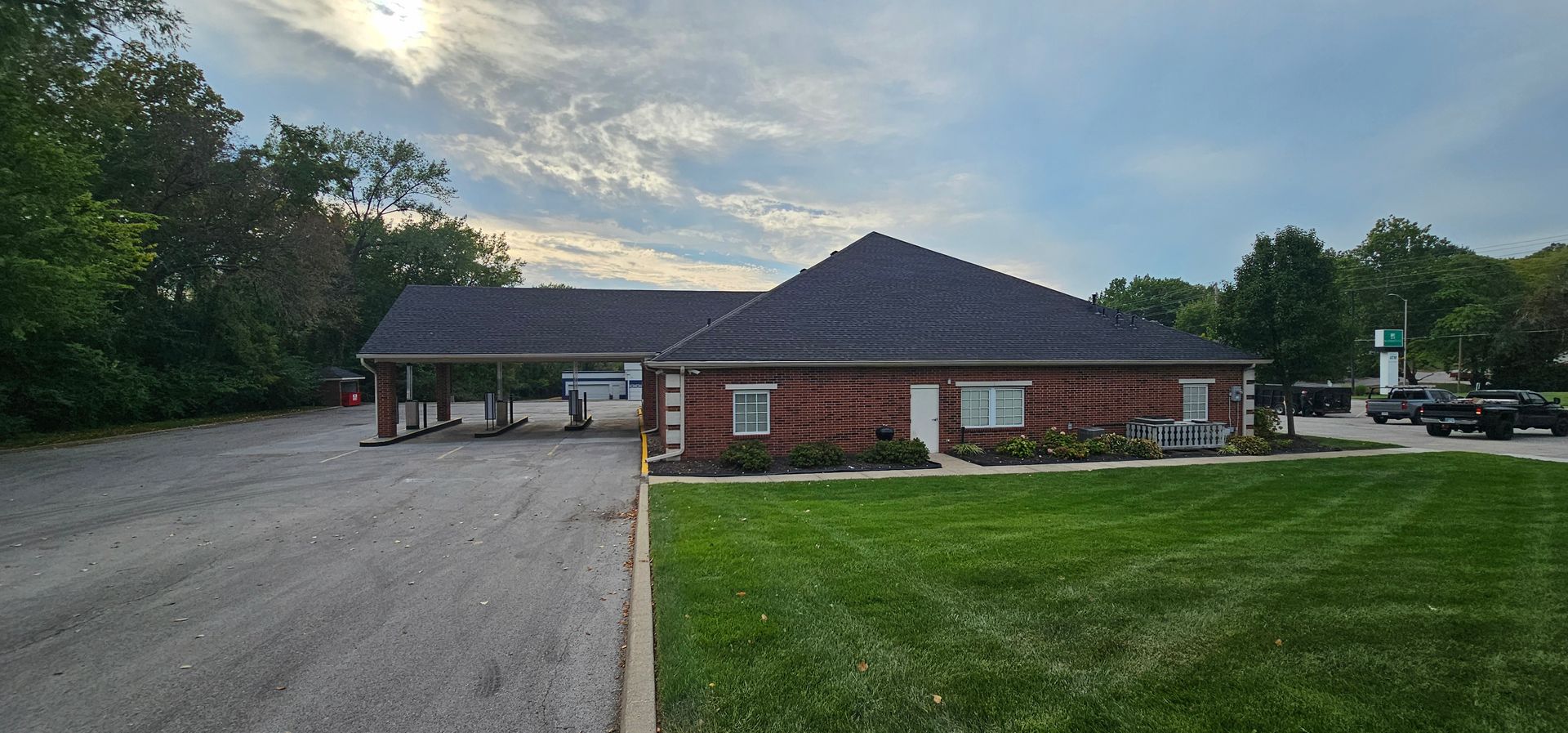Small brick building with a dark roof beside a gravel lot and green lawn at dusk