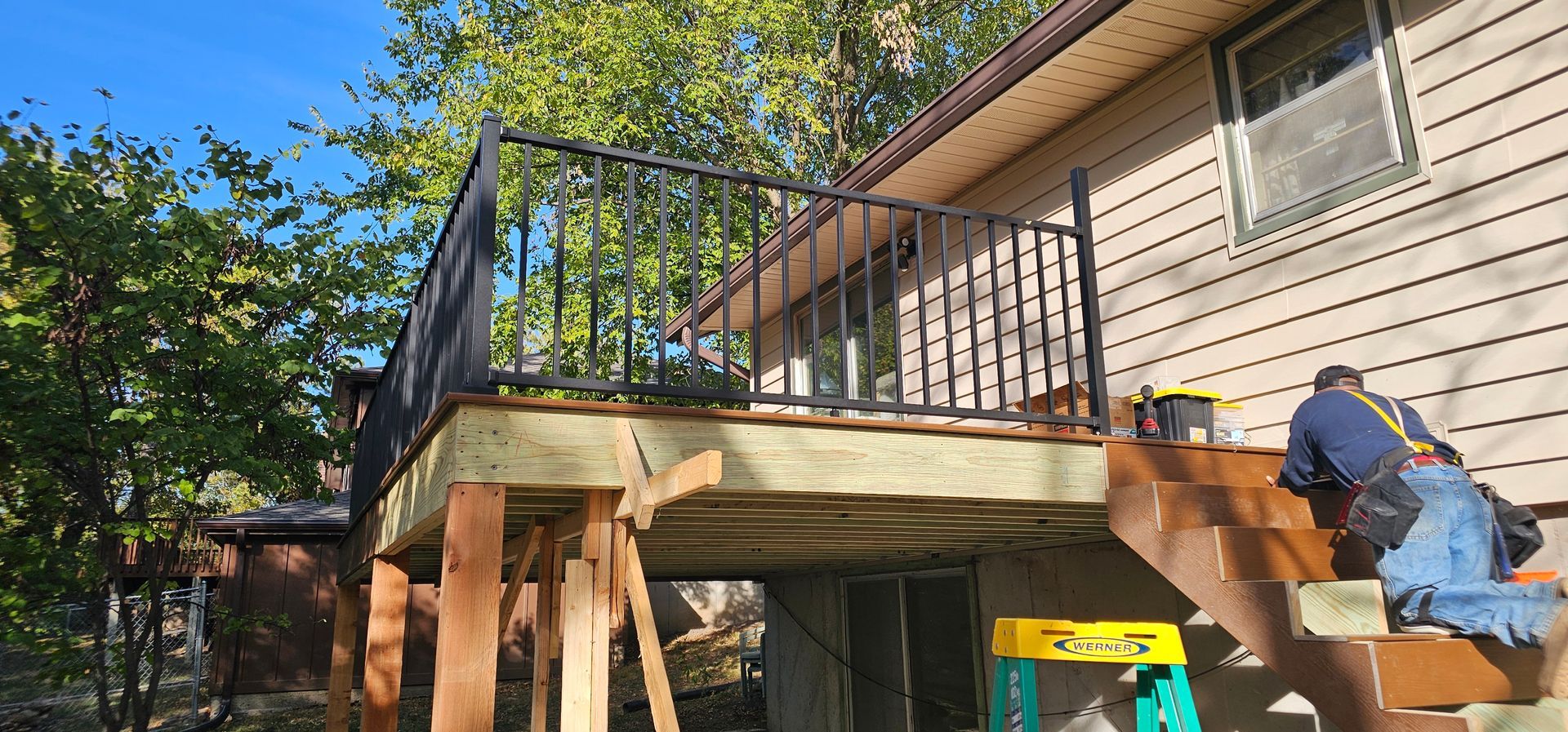 Worker sanding the side of a raised deck beside a tan house, with tools and black railings.
