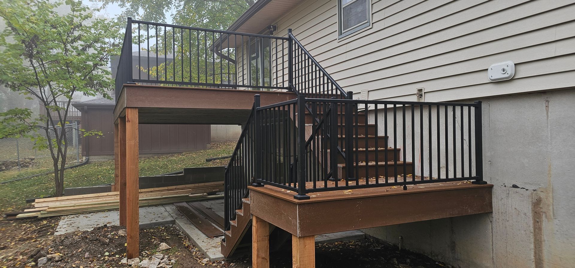 Raised wooden deck and stairs attached to the side of a house, with black railings and damp ground below.