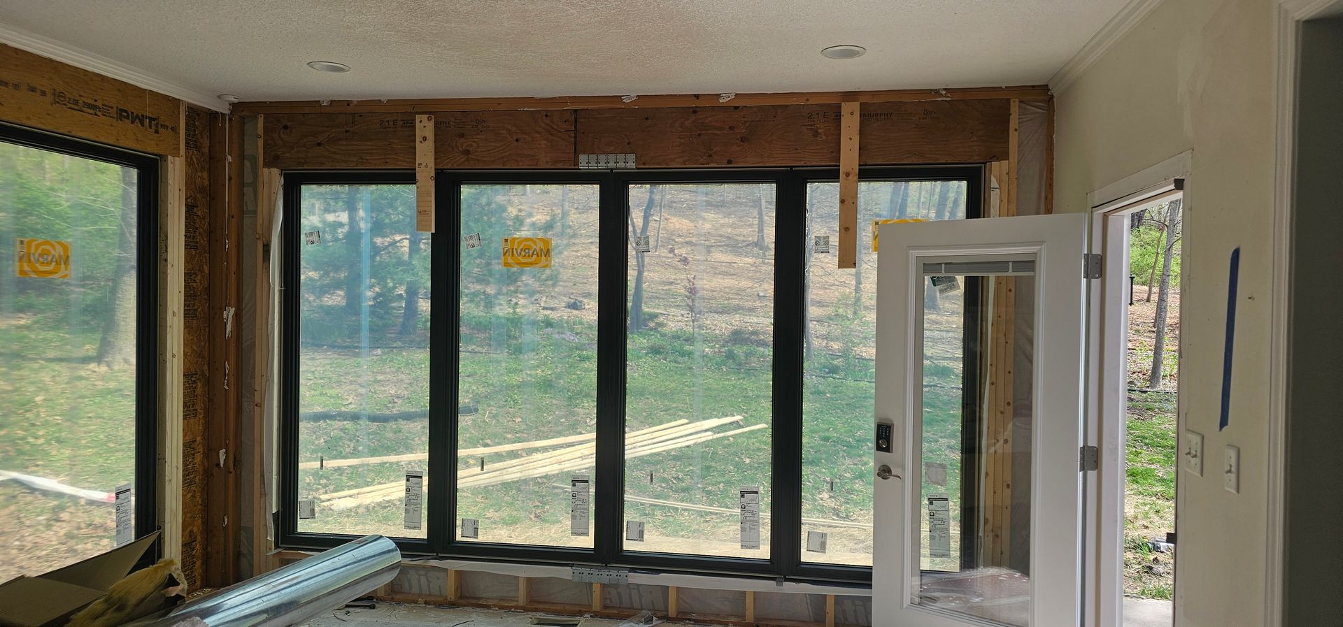 Empty room under renovation with large windows and an open white door, overlooking greenery outside