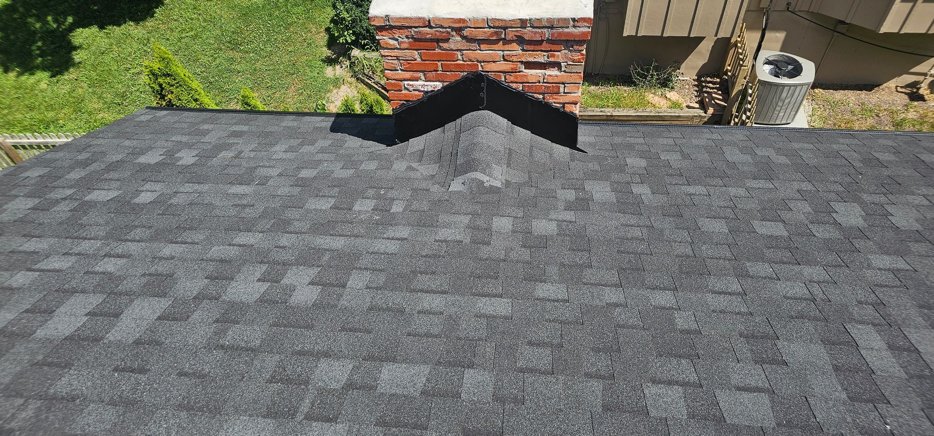 Gray shingle roof with a black vent near a brick wall and green trees in the background