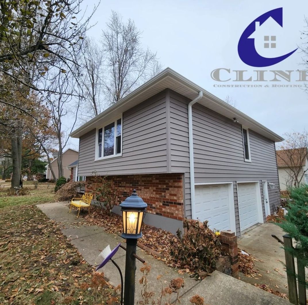 Suburban house exterior with gray siding, brick base, white garage doors, and a lit walkway lamp