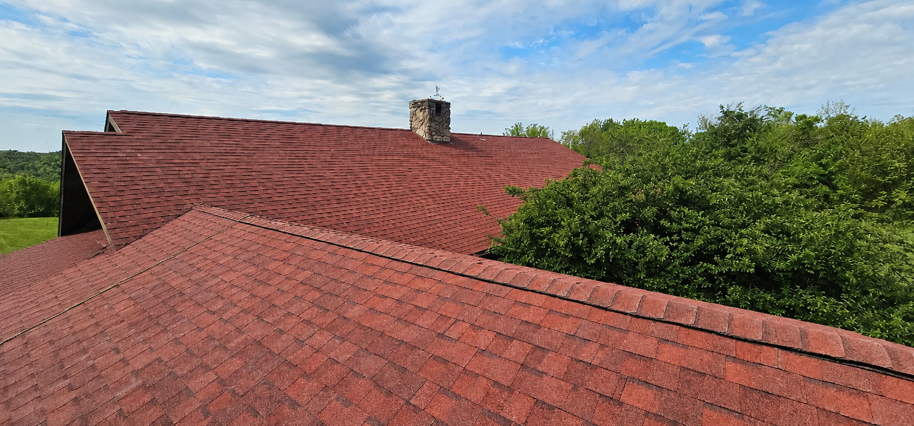 Red shingled rooftops with a chimney, beside green trees under a blue sky