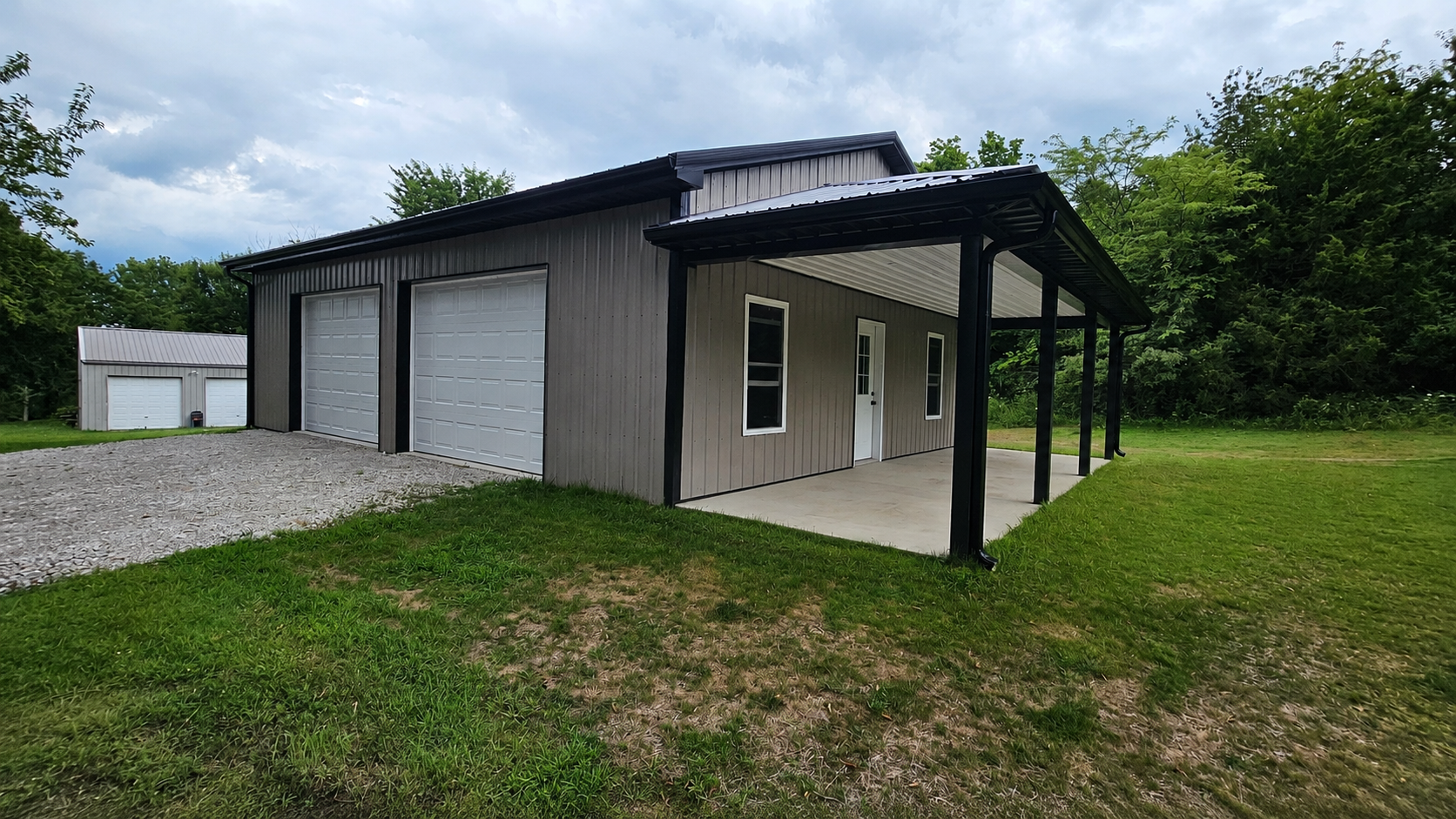 Small gray metal garage with porch on a grassy lot, under cloudy sky, beside a gravel drive