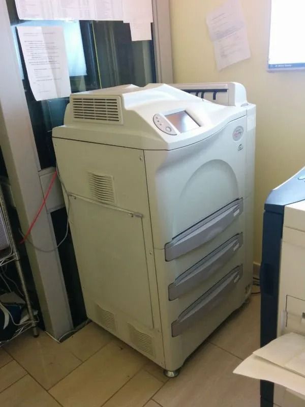 Una grande fotocopiatrice/stampante beige da ufficio in uno spazio di lavoro disordinato, accanto a una finestra e a una scrivania.