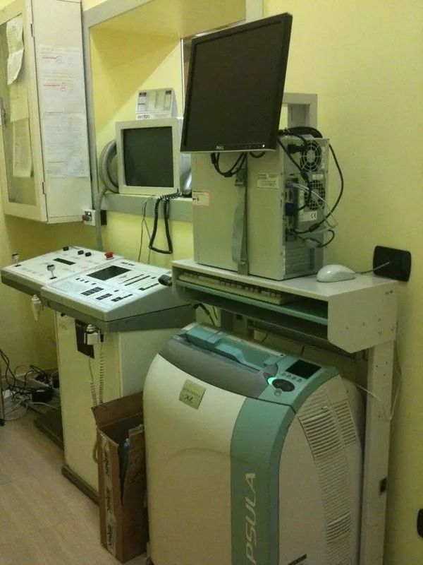 Postazione di lavoro per diagnostica per immagini medicali con monitor, pannello di controllo e apparecchiatura di scansione in una stanza stretta.