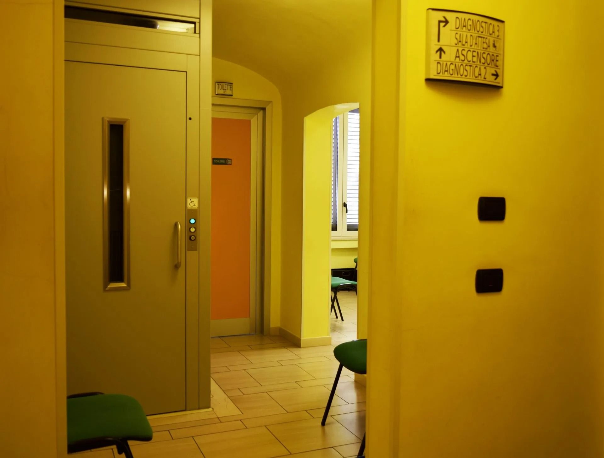 Corridoio giallo con ascensore, porta arancione, orologio a muro e sedie verdi.