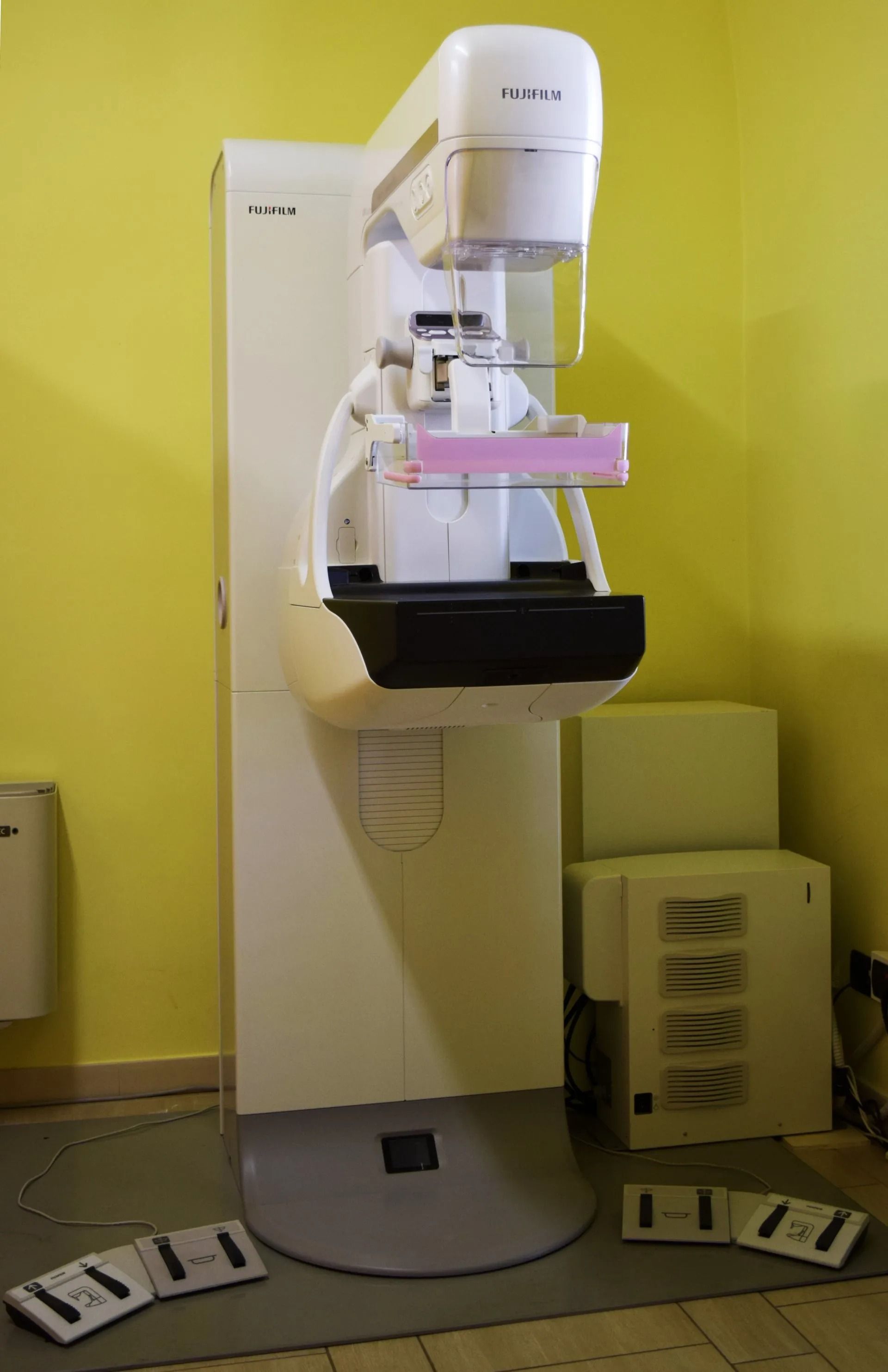 Apparecchio per mammografia in una stanza clinica con pareti gialle, posizionato su un piedistallo con vassoi nelle vicinanze.