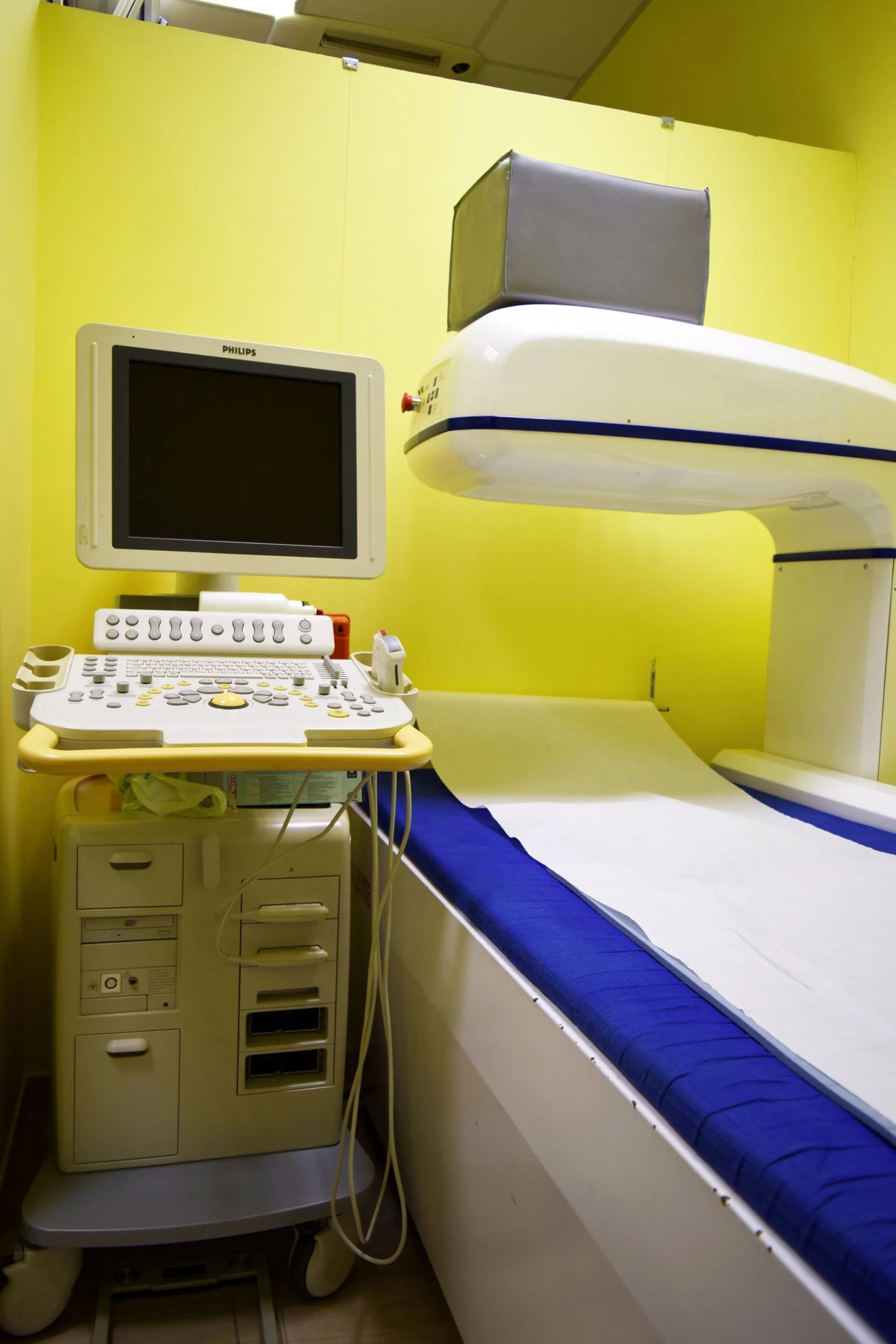 Sala visite ospedaliera con pareti gialle, monitor medico e lettino da visita con copertura blu.
