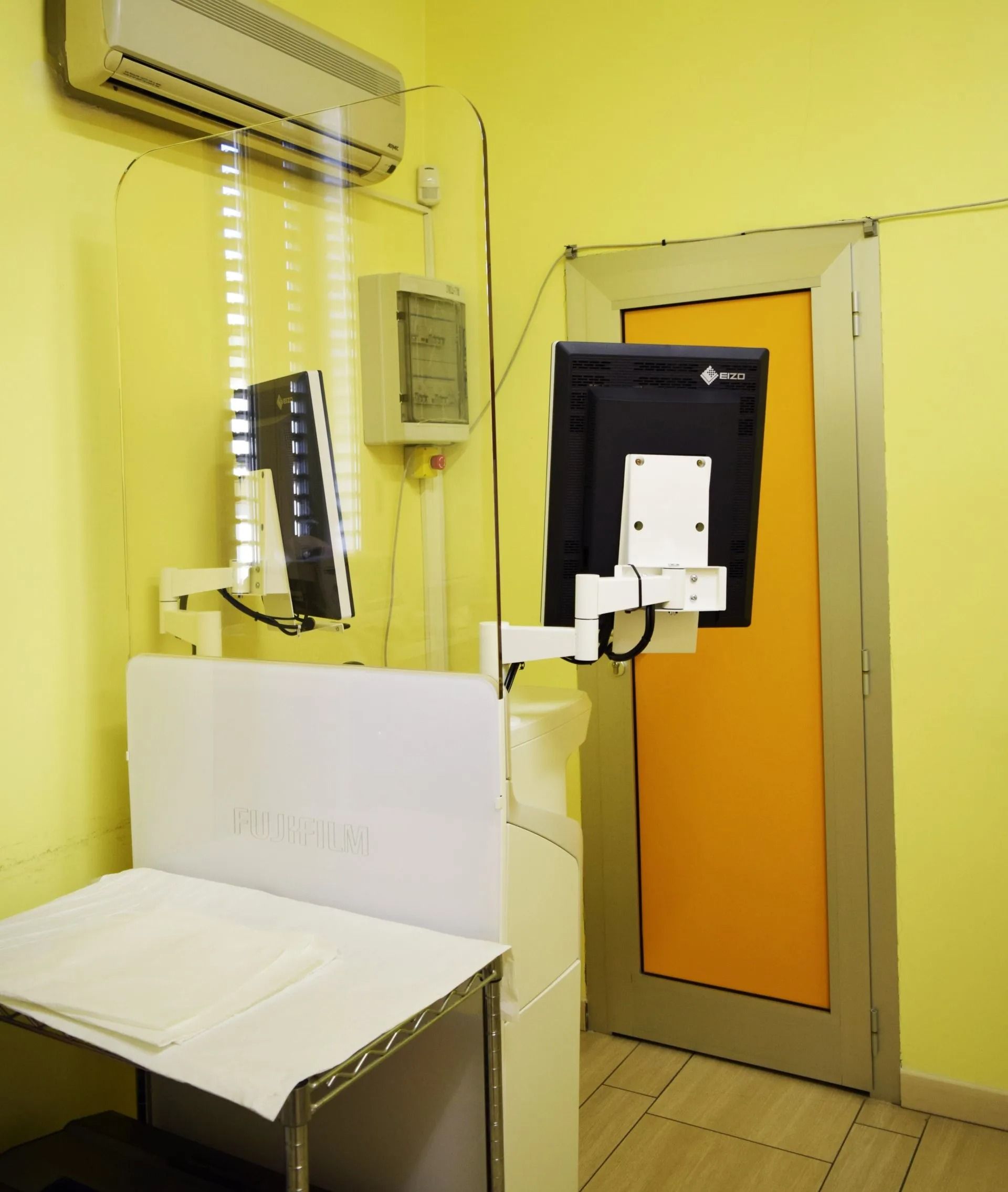 Piccola stanza con un letto bianco, condizionatore a parete e una porta con un pannello nero accanto.
