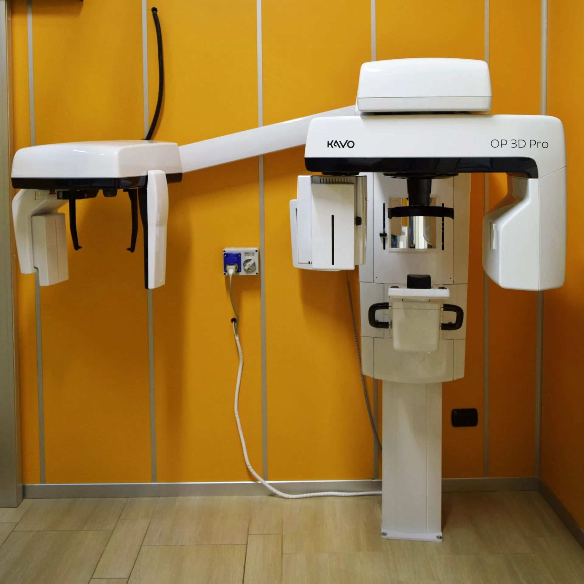 Apparecchio radiografico dentale in una clinica con pareti arancioni e pavimento in legno, che mostra due bracci scanner bianchi.