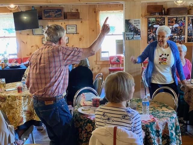 People dancing in a decorated room, man pointing up, woman with arms raised, others seated at tables.