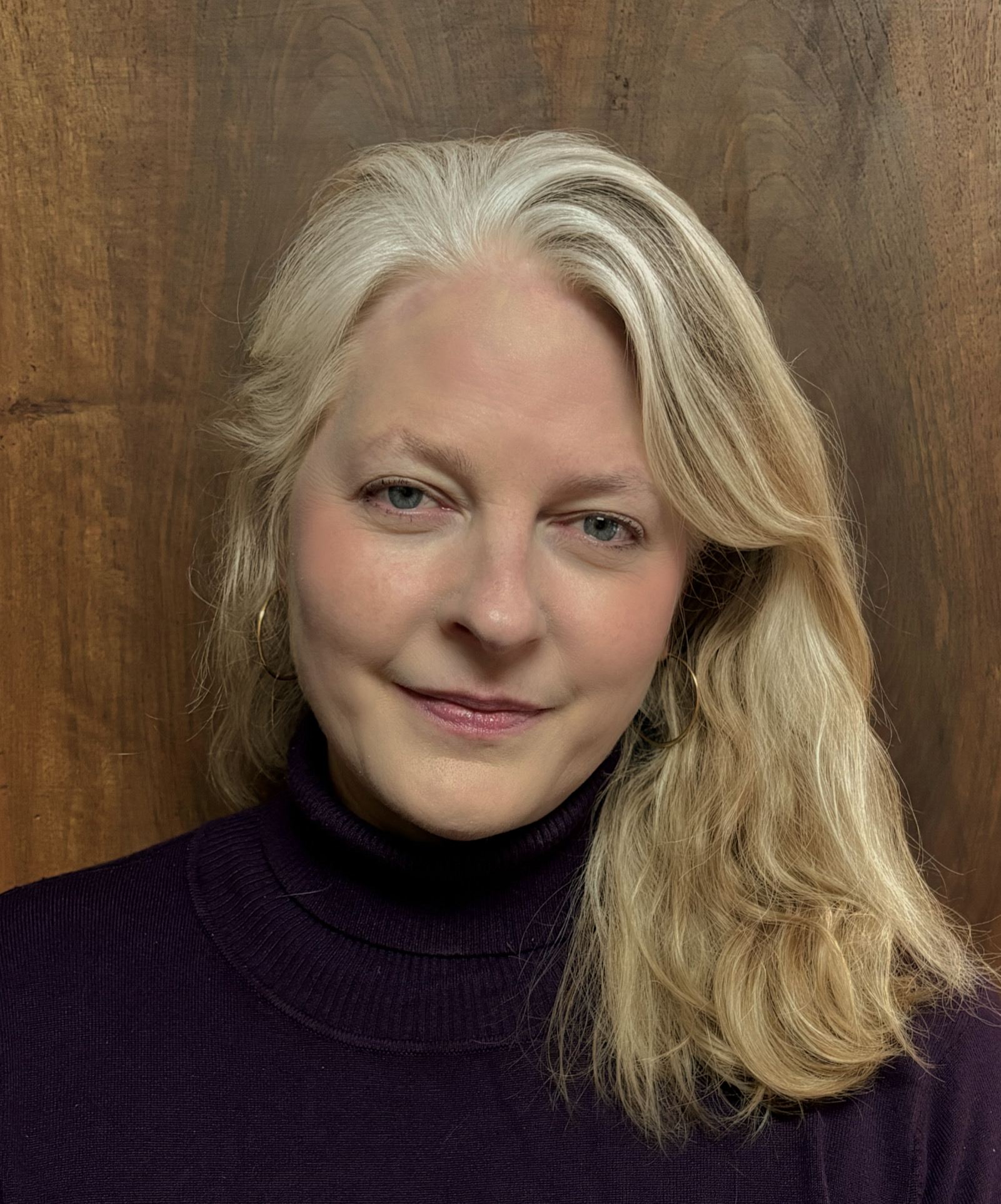 Woman with long, wavy blonde hair, wearing a purple turtleneck, smiling slightly. Against a wood-paneled background.