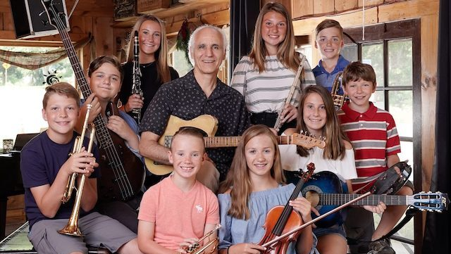 A group of musicians, adults, and children, inside a wooden building. Some hold instruments, smiling.