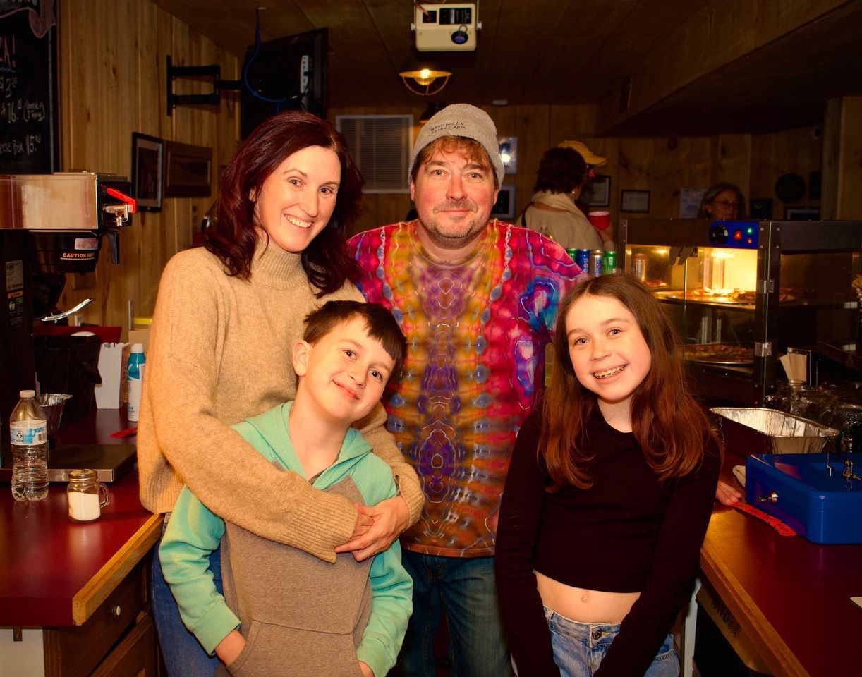Family smiling together inside a diner; woman in sweater, man in tie-dye, two children; red counter.